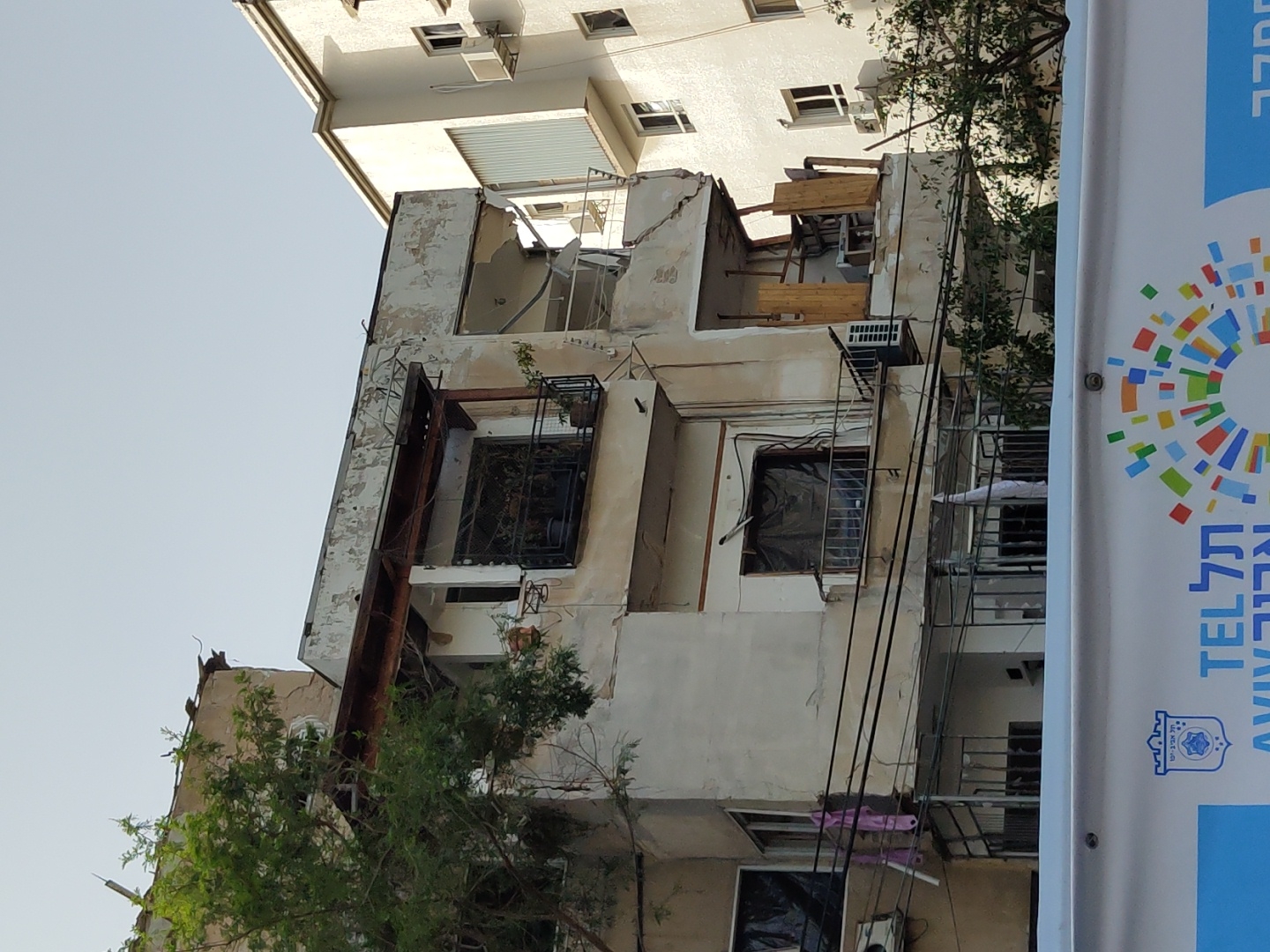 The image depicts a dilapidated building in Tel Aviv, Israel. The building shows signs of damage and decay, with cracked and crumbling facade. Visible are windows, some boarded up, and structural supports are in place. A banner with the words "TEL AVIV" in Hebrew and English, along with a city logo, is in the foreground. Trees are partially obscuring the building. Overhead wires are visible. The sky is clear.