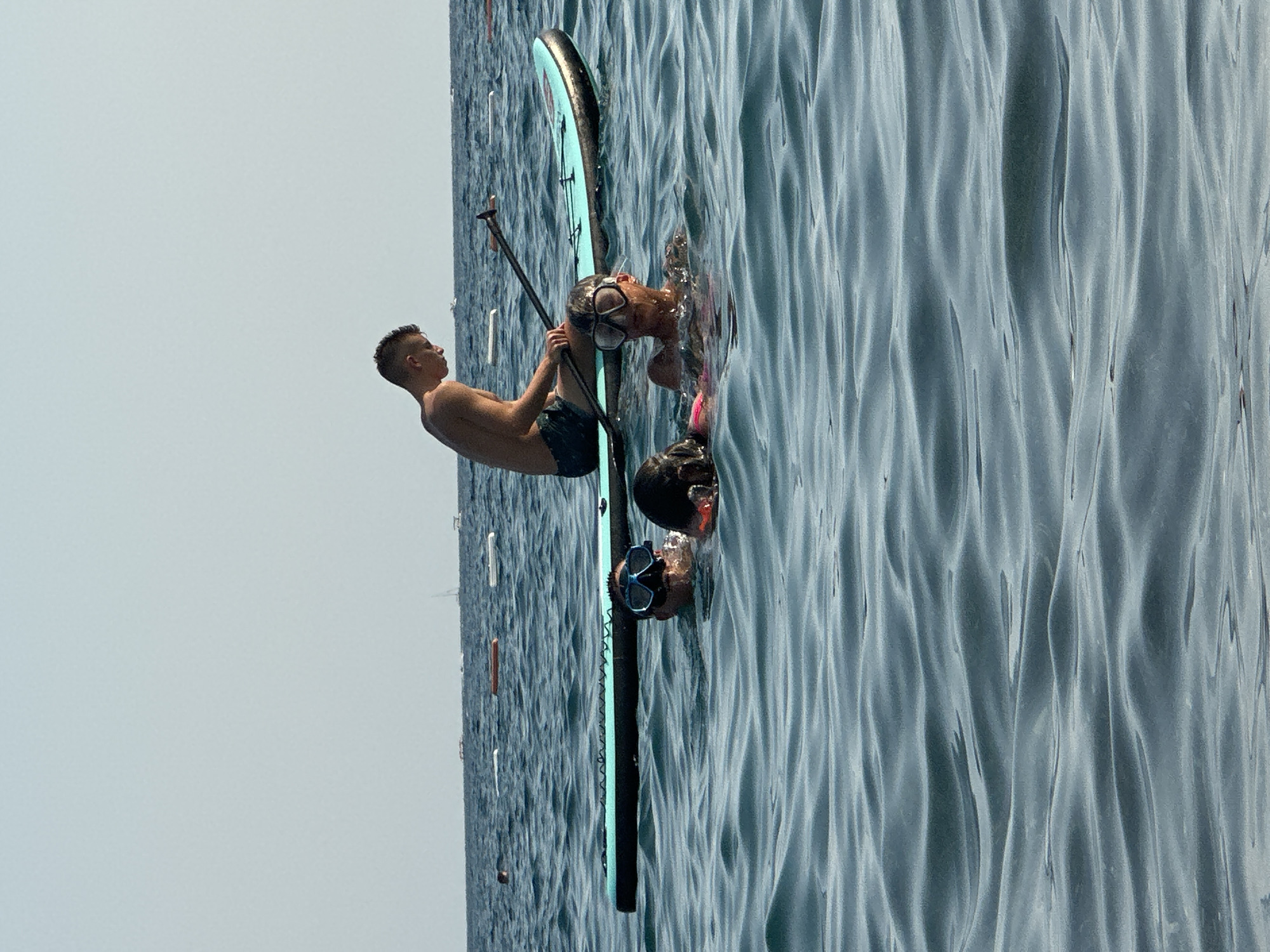 In the waters off Boreti, Montenegro, a group of people are enjoying recreational activities. A teenage male, wearing dark swim trunks, is seated on a turquoise paddleboard, holding a paddle. In the water surrounding the paddleboard, three individuals are visible. One person, wearing a diving mask, is looking towards the camera. Another individual, also wearing a diving mask, has their face submerged in the water. A third person is partially submerged, wearing a pink top and dark hair. The sea is calm with gentle waves. In the distance, several buoys and a sailboat are visible on the horizon under a clear sky.