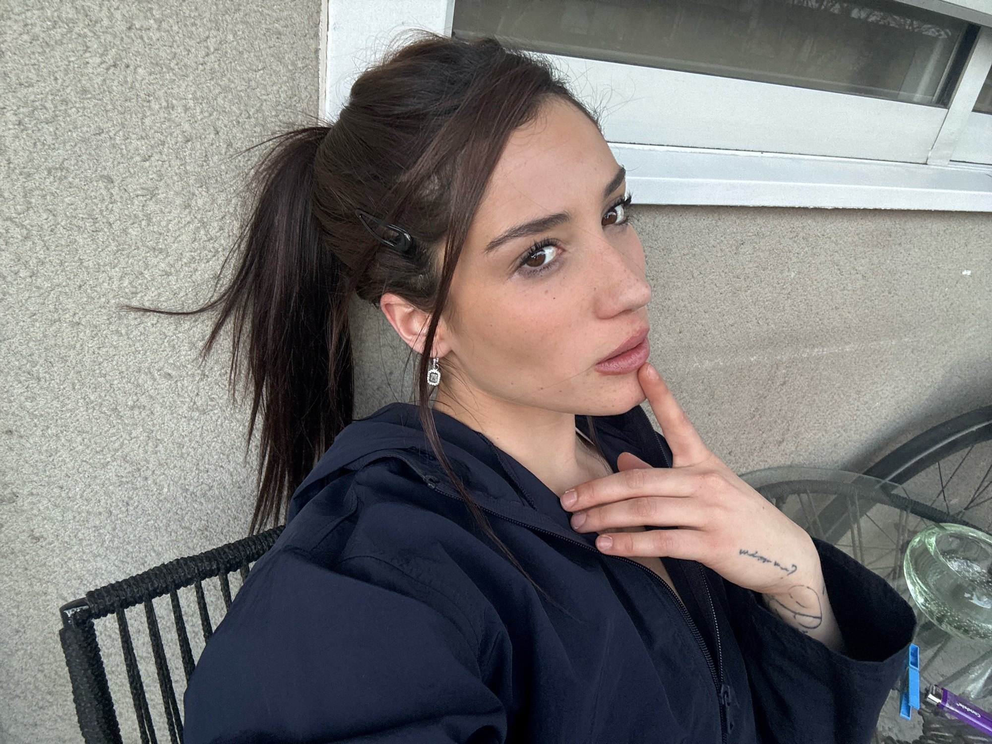 A young woman with brown hair pulled into a high ponytail is pictured from the chest up, seated on a black wicker chair. She wears a dark blue hooded jacket and a silver earring in her left ear. Her right hand is raised to her chin, and a tattoo is visible on her left forearm. The background consists of a textured, grey wall and a window with horizontal panes. To the right, a bicycle wheel and a glass table are partially visible. The scene appears to be an outdoor patio or balcony. The exact location in Buenos Aires, Argentina, cannot be determined from the visual information provided.