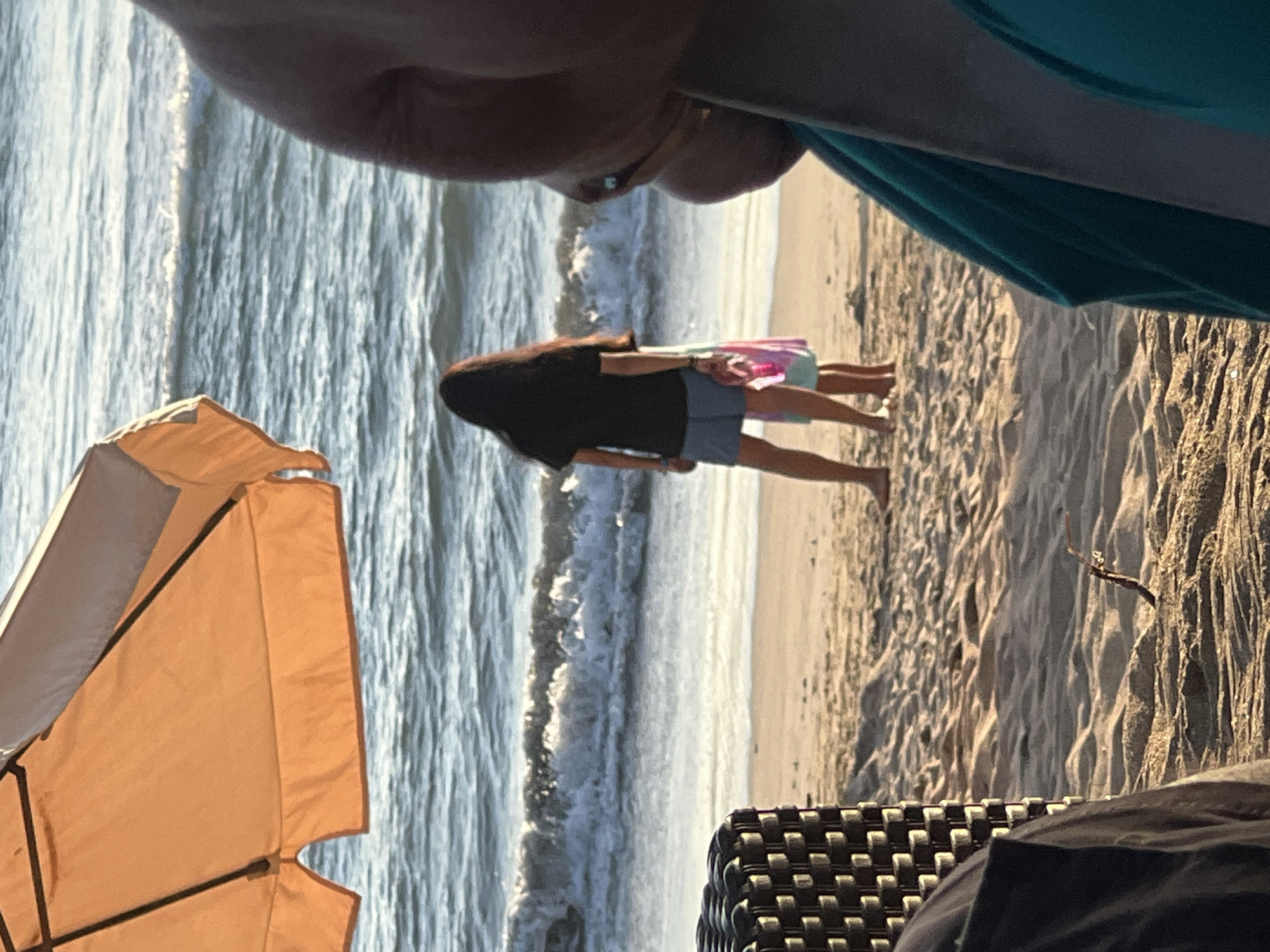 The image depicts a beach scene, likely Kuta, Indonesia, with soft golden sand and gentle ocean waves under a bright sky. In the foreground, part of an orange beach umbrella shades a dark woven chair and a person wearing a dark garment. Further back, two individuals stand near the water. One, facing away, has long dark hair and wears a black t-shirt and blue shorts, holding pink and blue flip-flops. The other person, partially obscured, appears to be younger, standing beside the first. The ocean, with its white foam and shimmering surface, creates a peaceful backdrop. A small piece of driftwood lies on the sand.