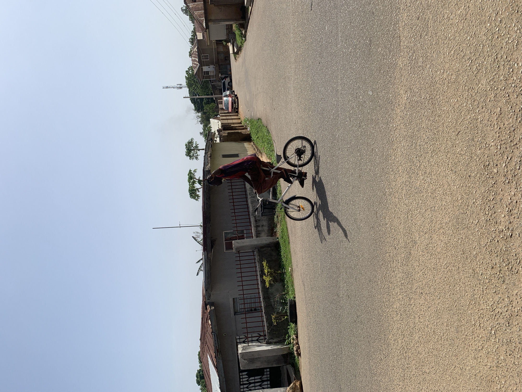 The image shows an individual riding a white bicycle on an asphalt road in what appears to be a residential area of Jos, Nigeria. The person is wearing a dark top and red pants. Buildings with fenced yards line one side of the road, while the other side is blurred, suggesting motion or focus. The sky is light blue with no clouds. Other vehicles are visible in the background. There is a cellular tower near the buildings.