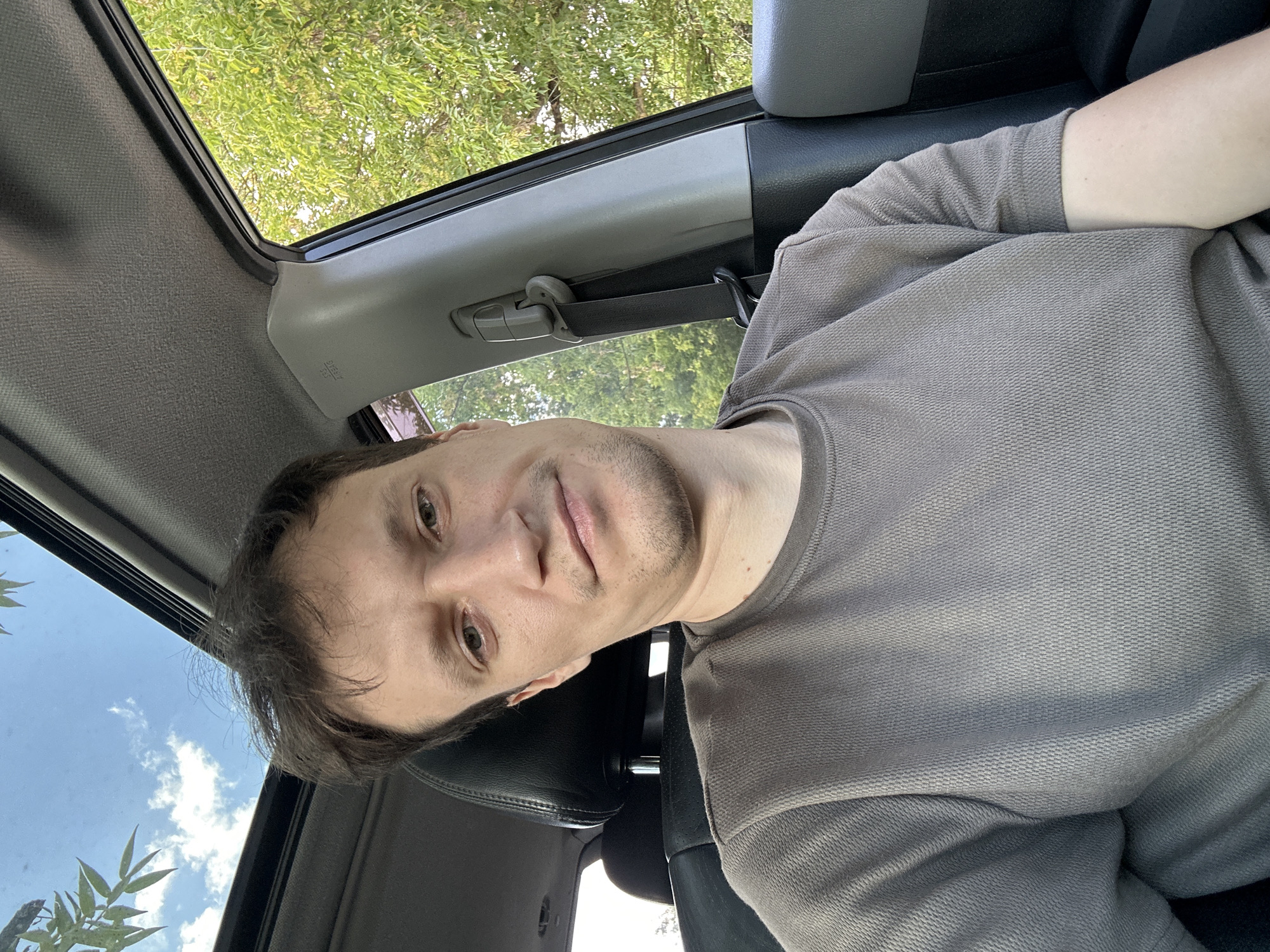 A young adult male with light brown hair and light-colored eyes is seated in the driver's seat of a vehicle. He is wearing a short-sleeved, gray t-shirt. The vehicle's sunroof is open, allowing a view of the sky and trees outside. The interior of the car is visible, including the car seat, headrest, and a portion of the car's interior paneling. The scene appears to be outdoors in a location with foliage, suggesting it is not within the urban core of Makiv, Ukraine, but rather on the outskirts or in a rural setting. The lighting indicates it is daytime.