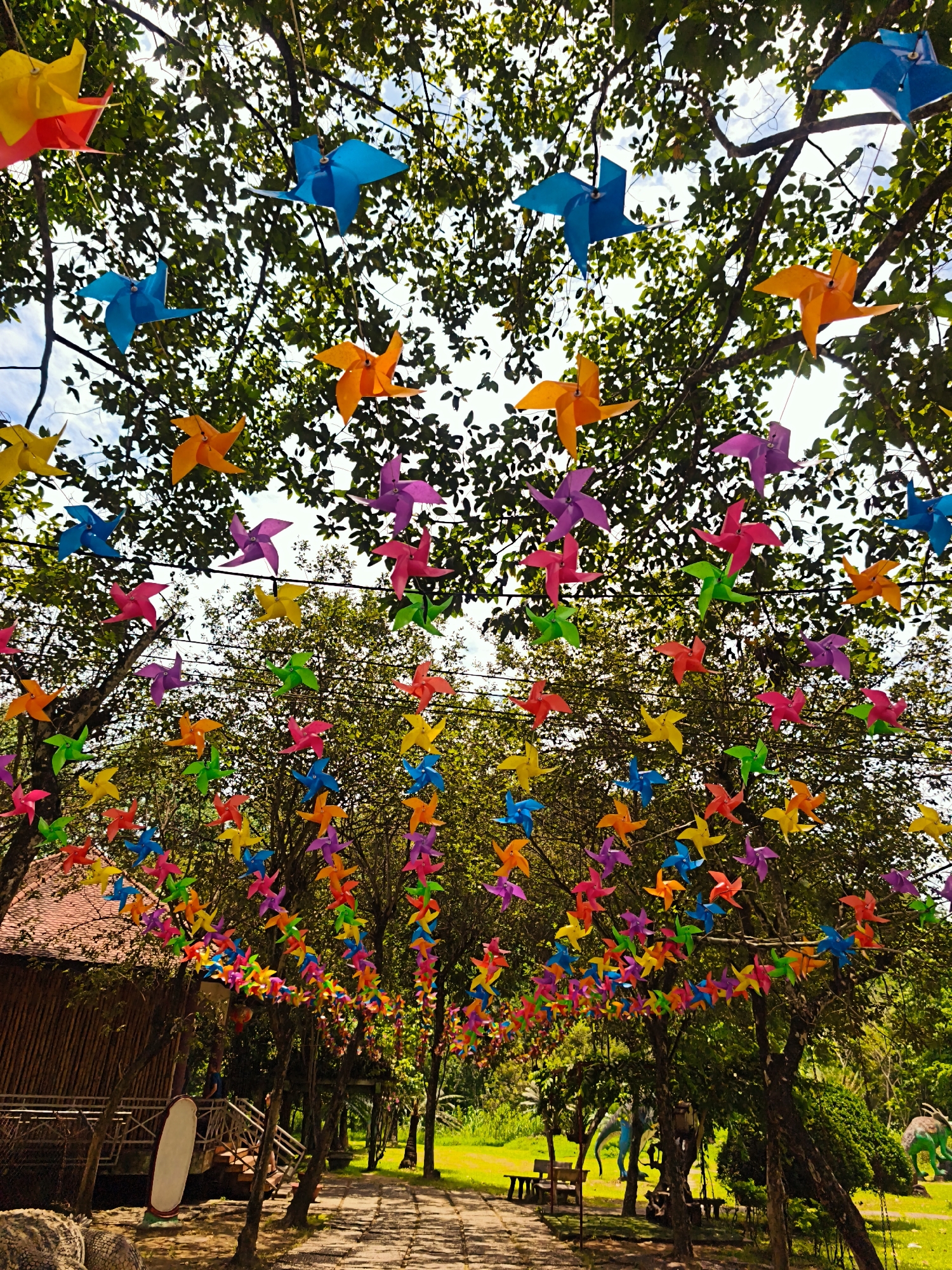 The image depicts an outdoor scene with numerous colorful pinwheels strung between trees. The pinwheels are in various colors, including red, orange, yellow, blue, green, and purple. They are suspended at different heights and form decorative garlands.
The trees are lush with green leaves, and the sky above is partly cloudy. In the background, there is a path leading through the area, and on the left side, a rustic wooden building with a reddish-brown roof is visible. Beyond the path, there are more trees, greenery, and some large, dinosaur-like sculptures. There are also benches and tables placed in the grassy areas.
The overall impression is of a festive or decorated outdoor space, likely a park or recreational area. The presence of the dinosaur sculptures suggests it might be a theme park or a children's attraction.
The prompt requests a location in Khanh Vinh, Vietnam, but there are no specific visual cues in the image to confirm this location. The scene is a generic outdoor setting that could be found in many tropical or subtropical regions.