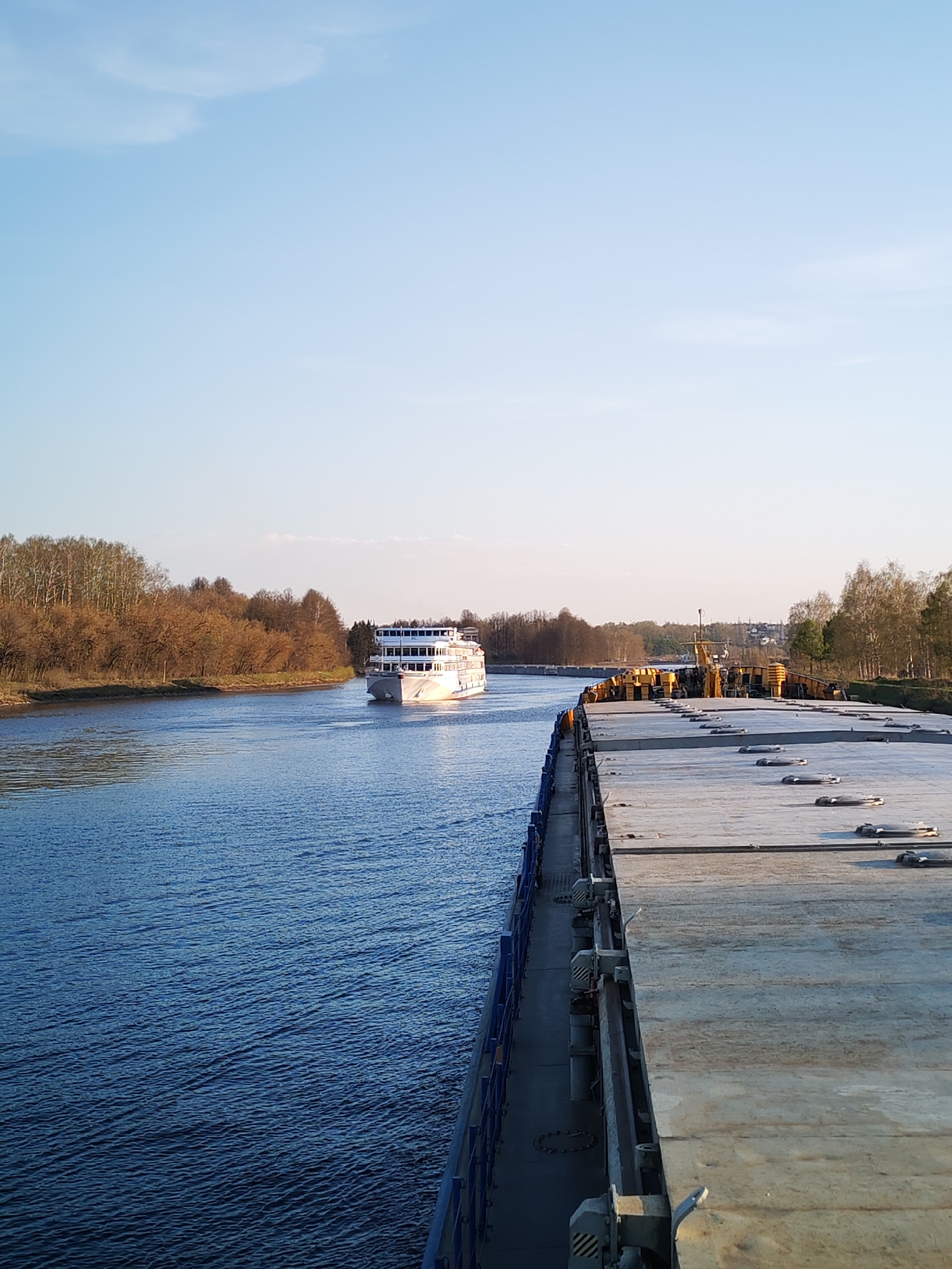 A sunny day in Базарово, Russia, features a river landscape where a multi-tiered white passenger ship navigates the waterway. The viewer's perspective is from the deck of a barge. The barge's gray, flat surface with circular hatches is prominent. To the left, the blue water of the river is bordered by trees with brown foliage. In the distance, there is another line of trees on the opposite bank. The sky is clear blue with a few faint clouds.