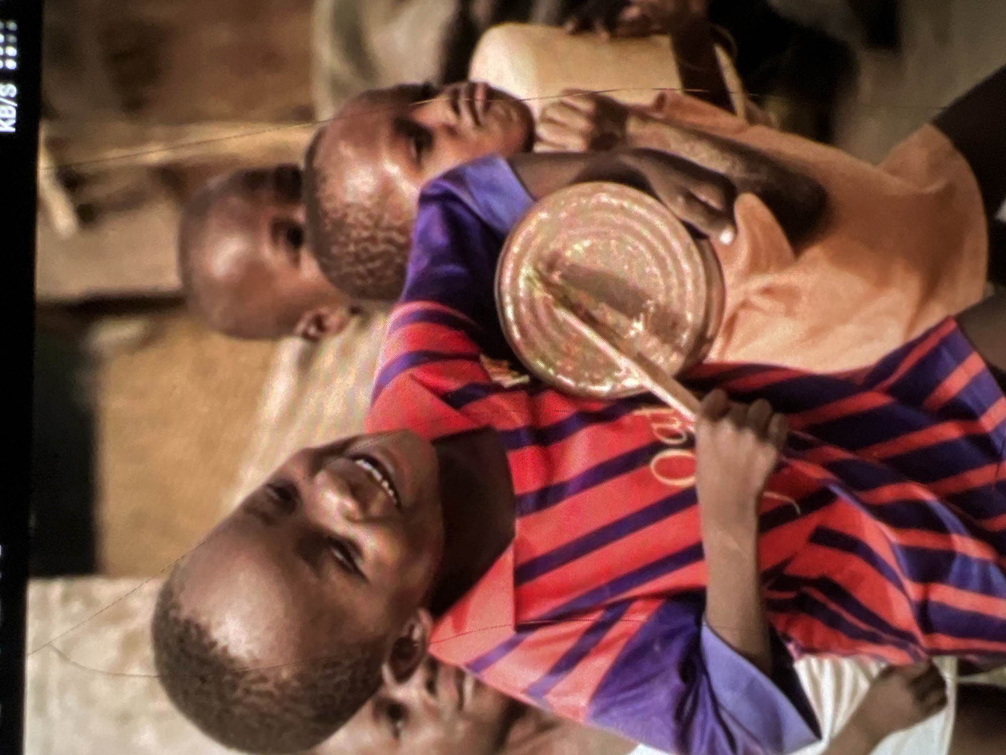 In a close-up, vertical shot, two young Black children are prominently featured, with other children partially visible in the background. The child in the foreground, facing left, is laughing with their mouth open, displaying teeth. They are wearing a red and purple striped shirt with a collar. Their right hand holds a round, metallic object with ridges, possibly a tin can, and their left hand is partially visible. Behind and to the right of the foreground child, another child is visible, facing slightly away, with their mouth pursed. They are wearing an orange garment. The background is blurred, suggesting an outdoor environment with a brown, earthy tone, possibly indicating a street or dusty area in Lagos, Nigeria. The lighting appears to be natural daylight.