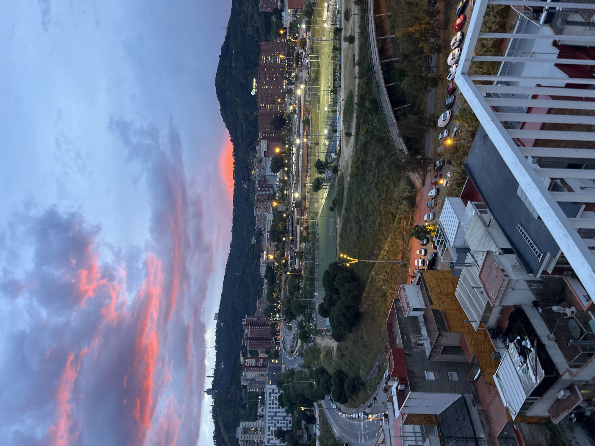 The image shows a cityscape at dusk, potentially in Barcelona, Spain, given the presence of buildings and a hillside. The sky features clouds with hints of pink and orange from the setting sun. A tree-covered hill runs vertically along the left side of the frame, with residential buildings scattered along its base and higher up. Streetlights illuminate roads and buildings. In the central area, there's a sports complex with lit fields. On the right, a balcony railing provides a viewpoint over rooftops and parked cars. The general atmosphere suggests an urban environment transitioning from day to night.