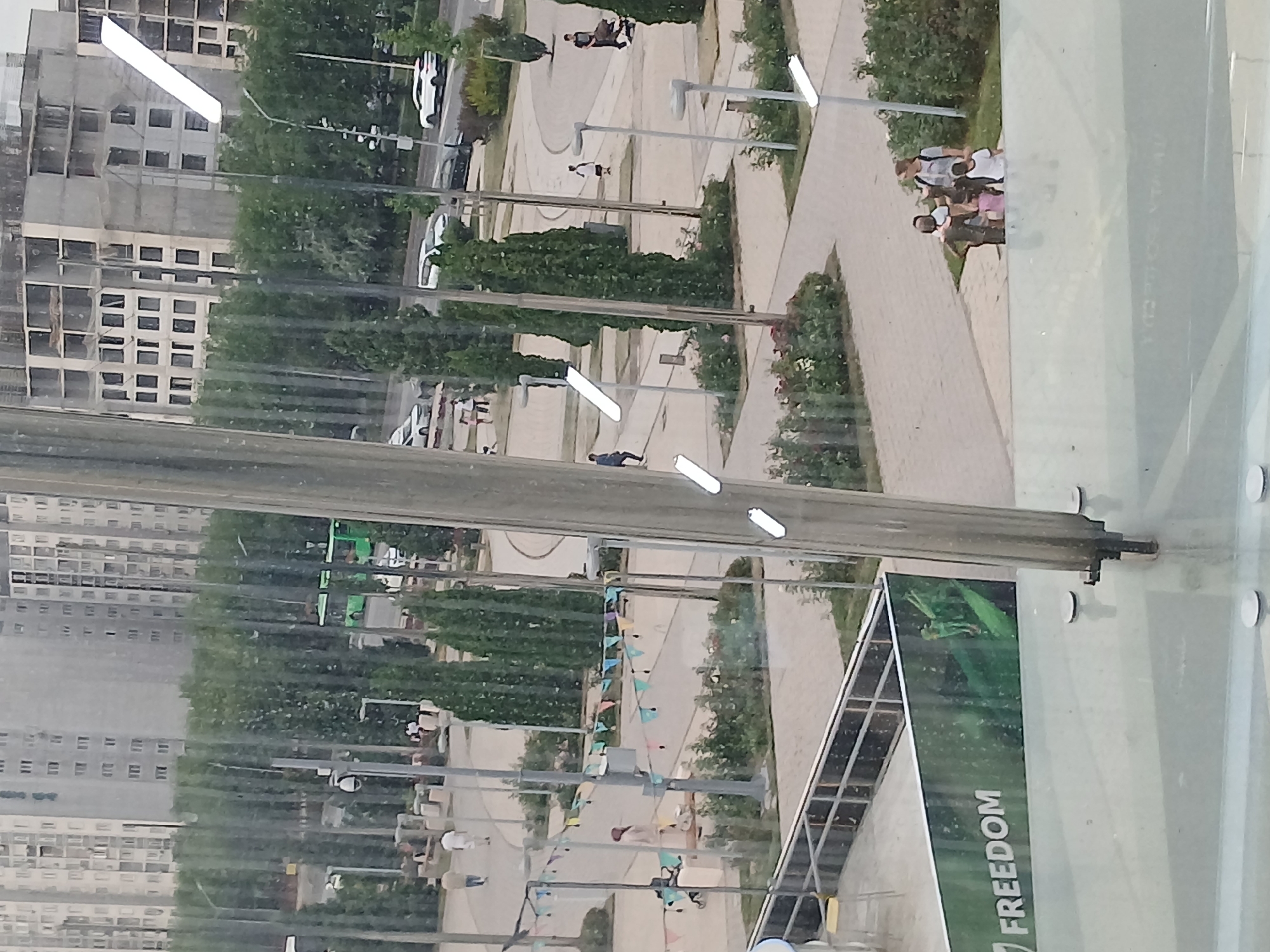 The image captures a blurred, daylight scene of an urban park or public space. In the foreground, a large, metallic cylindrical structure dominates the center, with several white, elongated light fixtures attached to it at varying heights. To the left and slightly behind this structure, a green banner with the word "FREEDOM" in white block lettering is visible, along with a graphic of green leaves. Beyond this, a metal railing and walkway are seen.
The midground reveals a paved area with pathways, dotted with numerous trees, many of which are slender and cone-shaped evergreens. Several people are present, including a group of children and an adult woman, and a man pushing a stroller. A person in lighter clothing is seen in the distance, and another individual walks alone on a paved path. A green bus is visible on a road in the background, along with several cars.
The background is filled with tall, multi-story apartment buildings, some appearing to be under construction with exposed frameworks. The sky is overcast, contributing to the diffused lighting of the scene. The overall impression is one of a public gathering space within a densely populated urban environment.