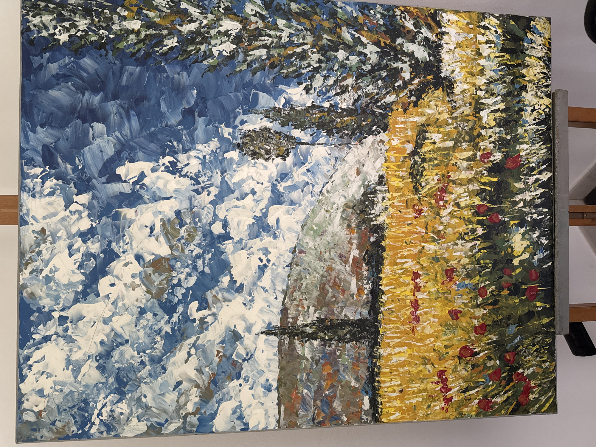 This is a painting of a landscape with a field of yellow flowers in the foreground, dotted with red flowers. In the mid-ground, there are several tall, dark trees, and behind them, a rolling hill with various colors suggesting foliage and earth. The sky is filled with dramatic, textured clouds in shades of blue and white. The style is impressionistic, with thick, visible brushstrokes. The painting is displayed on an easel. There are no people or any indication of the city of Cunardo, Italy.