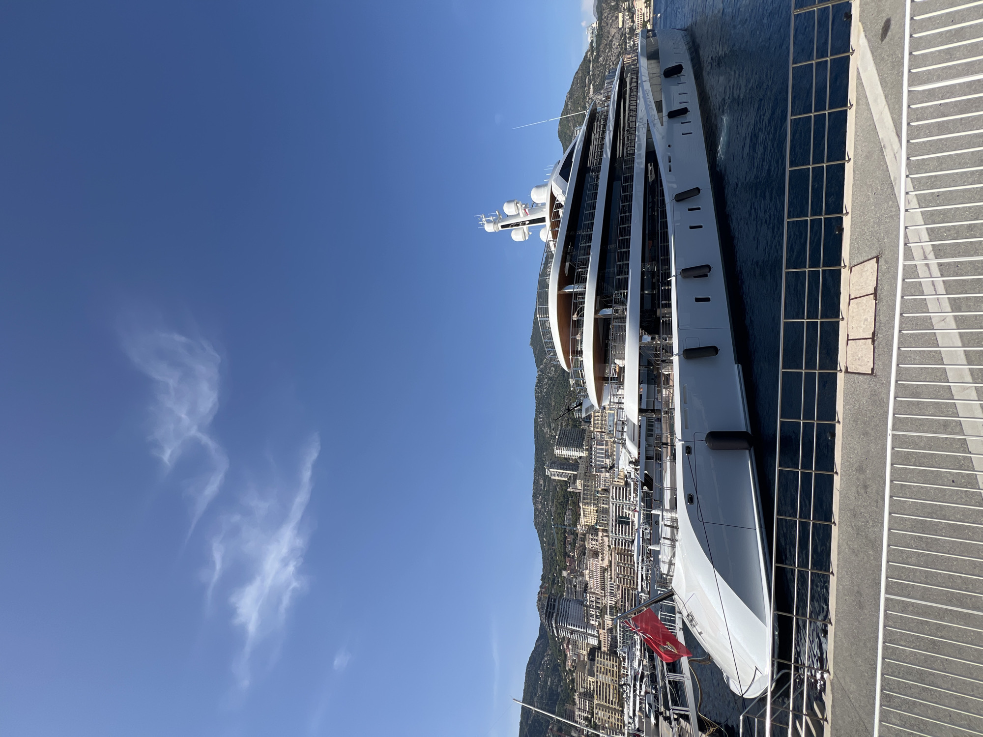 A wide-angle, eye-level, full shot captures a large, white, multi-deck superyacht docked in a harbor with the city of Monaco in the background. The superyacht is the primary subject, positioned on the right side of the frame and extending towards the left. It is white with multiple levels, featuring dark-tinted windows and balconies. A red flag with a crest is visible on the front left of the yacht. The water in the harbor is deep blue. Beyond the yacht, a dense, hilly cityscape of Monaco is visible, with numerous buildings of varying heights and architectural styles climbing up the slopes. The sky above is clear and bright blue, with a few wispy white clouds. A white metal railing runs along the bottom of the frame, separating the foreground from the harbor and the docked vessels.