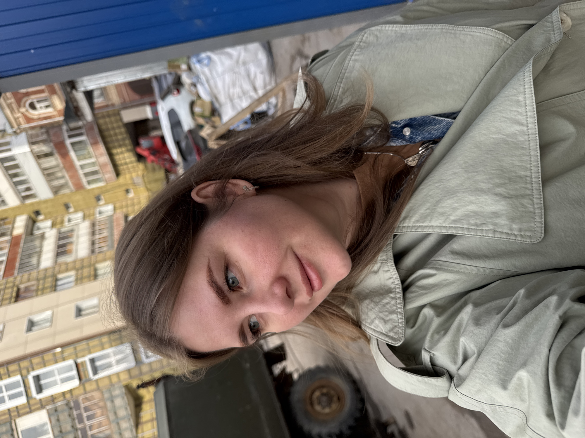 A woman is photographed from a high-angle perspective. She has fair skin, light brown hair, and blue eyes. She is wearing a light green trench coat over a blue denim shirt or jacket, and a silver necklace. She is looking slightly downward, toward the right of the frame. Behind her, there is a cityscape. Buildings with multiple windows and architectural details are visible, suggesting an urban area. A white car and other urban elements are also visible, although slightly out of focus. The presence of specific architectural styles in the buildings suggests the city of Perm, Russia. Also in the background is an olive green vehicle.