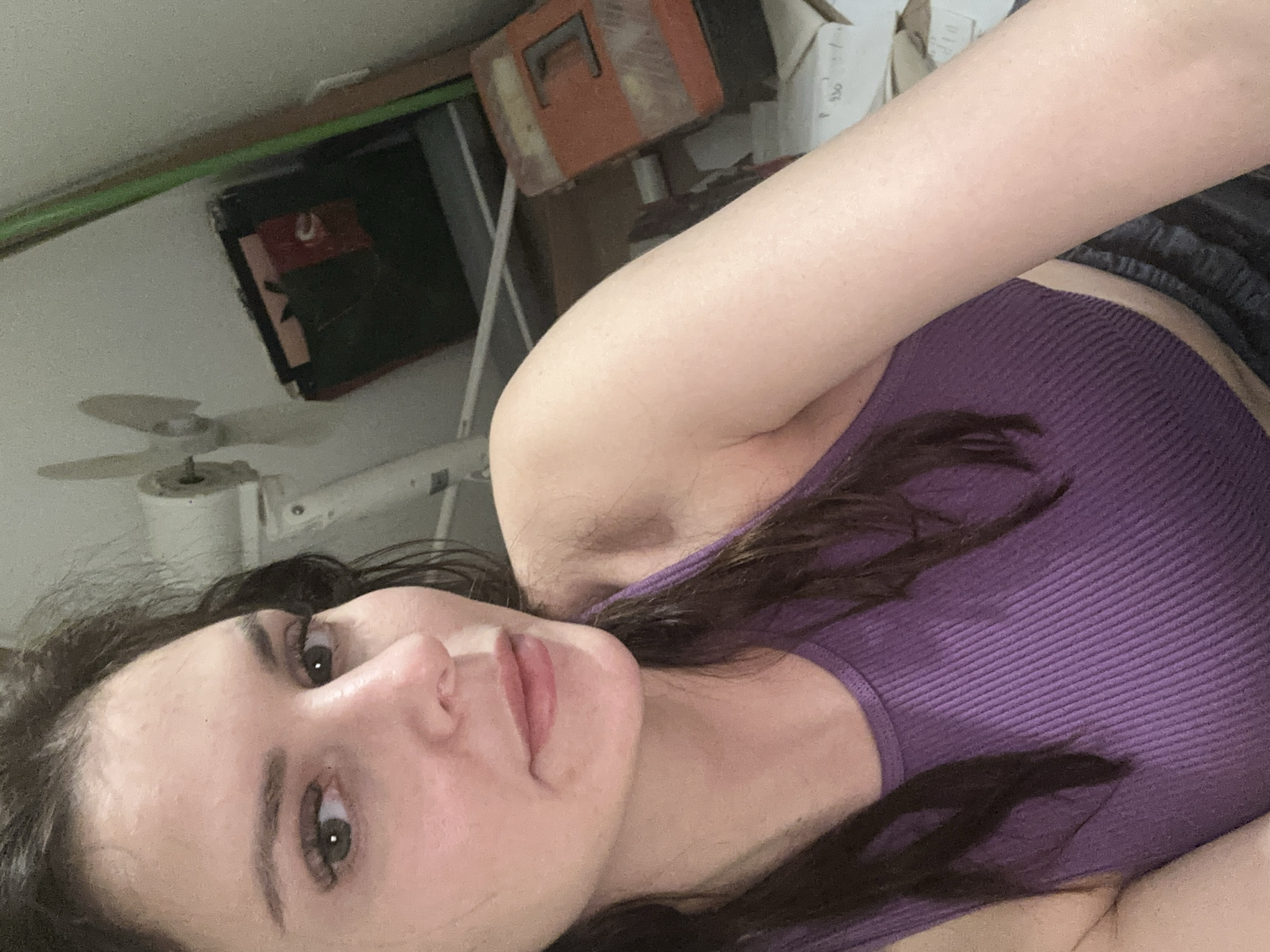 A young woman with dark hair and light skin is shown from the chest up in a selfie. She is wearing a purple ribbed tank top. Her makeup is visible, including mascara and lip gloss. In the background, there is a white ceiling fan, shelves with various items, and some cardboard boxes. The environment appears to be indoors, possibly a storage area or a room with clutter.