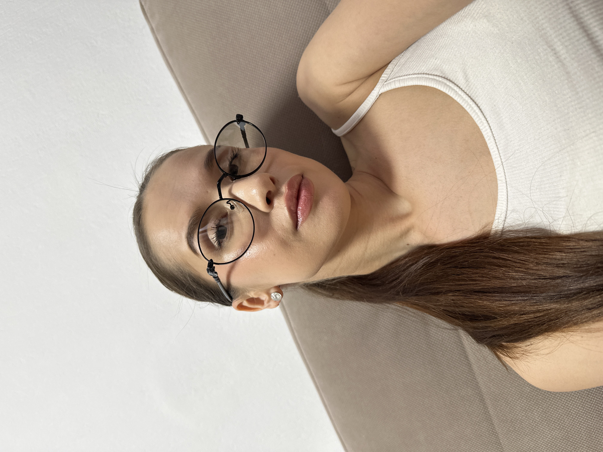 A vertical portrait features a young woman with long, dark brown hair and round, black-framed glasses, lying down and looking directly at the camera. The image is oriented such that she appears to be resting on her side, with her head towards the top of the frame. She is positioned on a light brown or beige textured sofa or bed, against a plain white or light-colored wall.
The woman, appearing to be in her 20s or 30s, has her hair pulled back, revealing her forehead. A small, clear stud earring is visible in her right ear. Her lips are full, adorned with a light pink or nude lipstick. She is wearing a light-colored, possibly white or off-white, ribbed tank top or camisole. Her gaze is direct and neutral, conveying a calm or contemplative emotion. The indoor lighting is soft and diffused, suggesting a daytime setting. There is no visible text in the image. While the provided context mentions Yekaterinburg, Russia, there are no specific visual cues within the photograph to indicate this location.