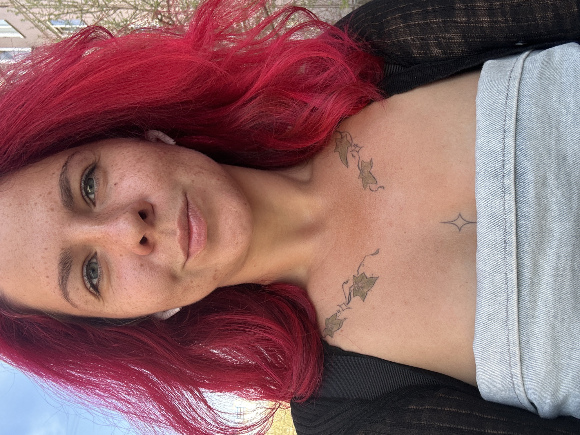 A young woman with striking, vibrant red hair is captured in a close-up portrait, looking directly at the camera. She has fair skin with numerous freckles across her nose and cheeks, and her eyes are a light green or hazel color. Her long, wavy red hair cascades around her head and shoulders.
She is wearing a sheer black top, and a light blue fabric, resembling denim, is visible on her left side. Notable details include an intricate tattoo of an ivy vine with green leaves extending across her right collarbone and shoulder area. A smaller, abstract star-like tattoo is also visible on her upper central chest. Small white earbuds are present in her ears. She maintains a calm, neutral expression with her head slightly tilted.
The setting appears to be outdoors during daylight, indicated by the soft, natural lighting. In the background, hints of green foliage, likely trees or bushes, and a patch of clear blue sky are visible. The image conveys a casual, personal moment. There is no visible text in the media. The location context for this photograph is Novosibirsk, Russia.
