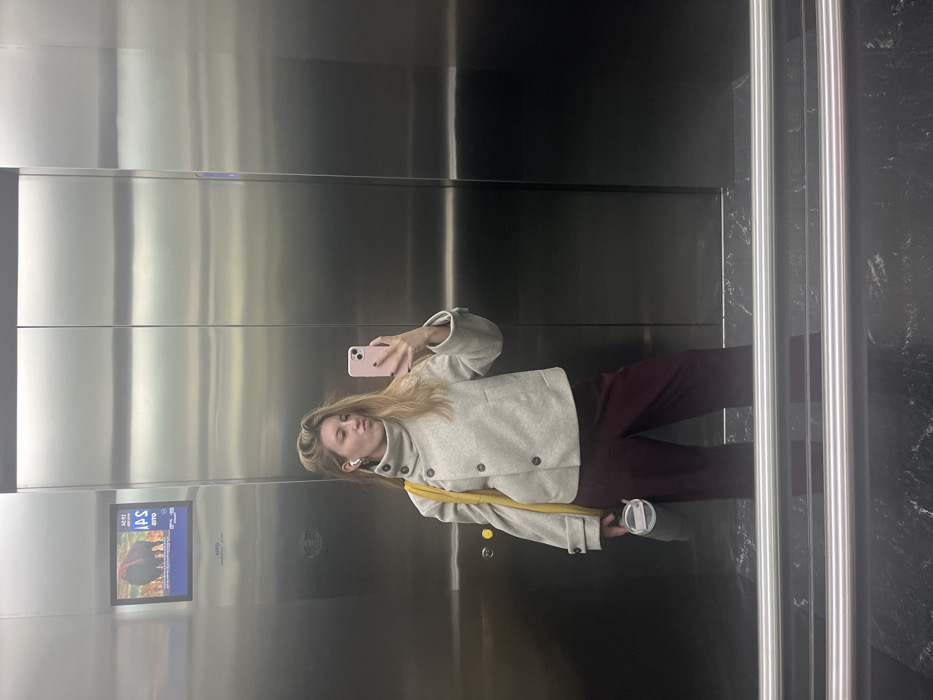 A woman takes a selfie in an elevator. She wears a light-colored coat, burgundy pants, and an earbud in her left ear. She holds a pink phone in her right hand and a to-go cup in her left. A yellow strap of a bag is slung across her shoulder. The elevator has stainless steel walls and a digital display. The elevator door is visible on the right.