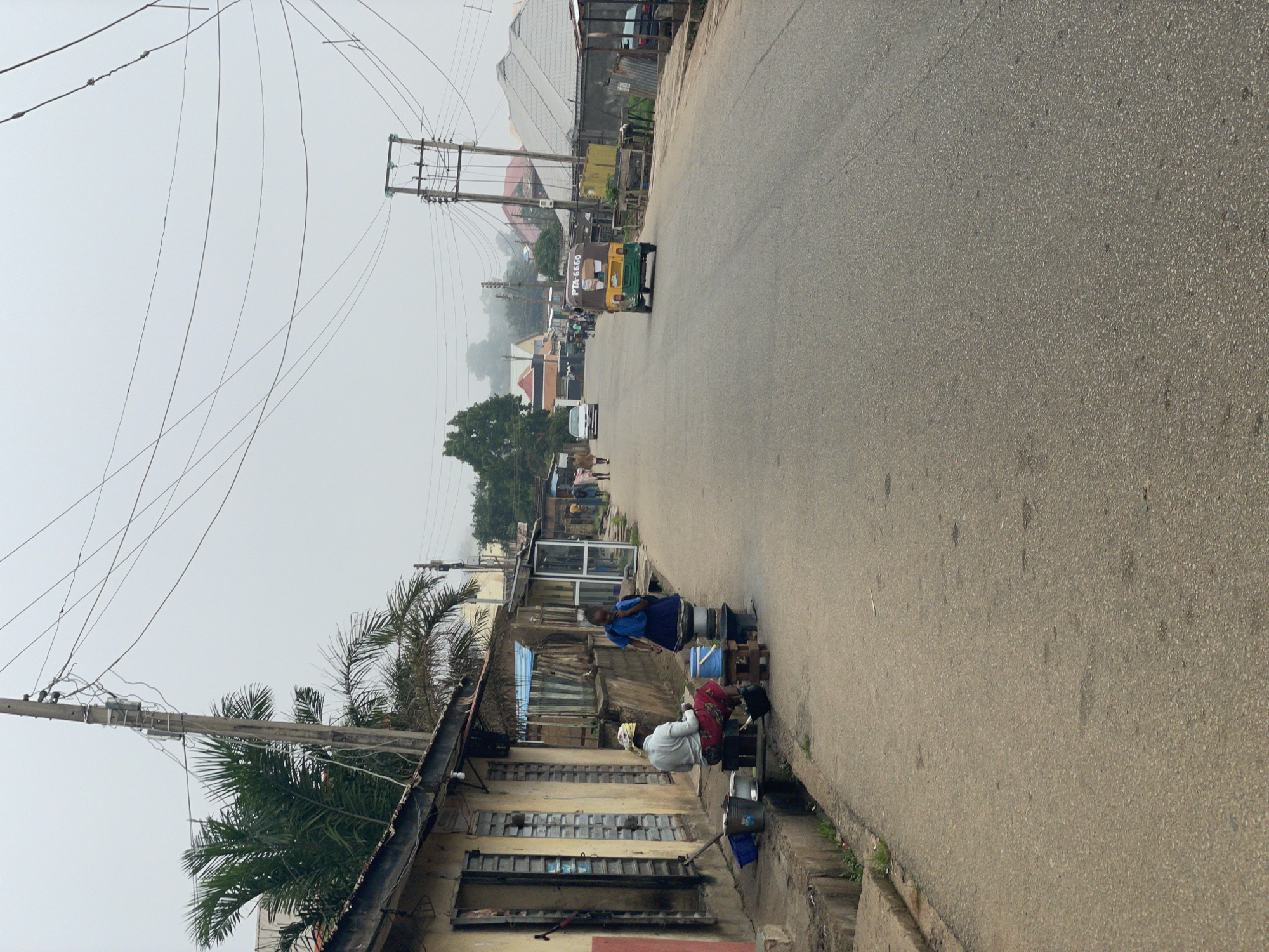 The image depicts a street scene in Jos, Nigeria. The street is paved with asphalt and appears to be a secondary road with some commercial establishments lining the left side. The buildings are single-story structures with open doorways and windows, some appearing to be shops. On the left side of the street, in the foreground, two individuals are visible. One person is seated on a stool, and another person is standing nearby. Several cooking pots and buckets are arranged on the ground in front of them, suggesting some form of street vending or food preparation.
Further down the street, a yellow and green three-wheeled motorized vehicle, commonly known as a Keke Napep, is driving away from the camera. There are other vehicles and pedestrians visible in the distance, indicating moderate activity on the street. Utility poles with numerous wires are a prominent feature in the background, typical of urban infrastructure. The sky is overcast, creating a diffused light that softens the scene. The overall environment suggests a developing urban area with a mix of commercial and residential structures.