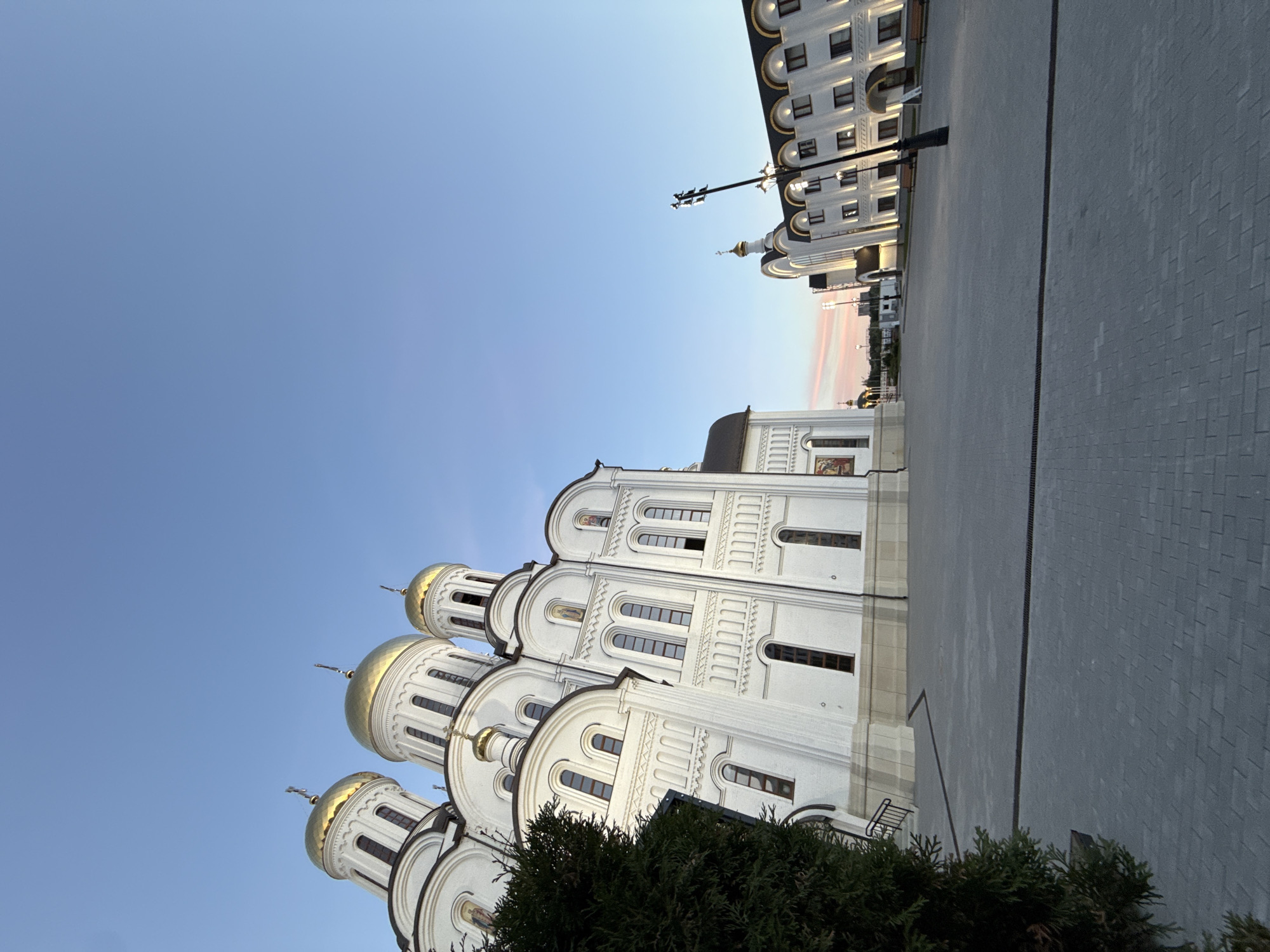 This is a vertical, eye-level, medium shot of a large, white Orthodox church with golden domes. The church is the main subject, viewed from a low angle, emphasizing its grandeur and multiple domes. To the right of the church, a modern, light-colored building with many windows is visible. The foreground features some dark green, coniferous trees. The background is a clear, pale blue sky, suggesting it is daytime. The light suggests either early morning or late afternoon, with soft light illuminating the scene. The ground in front of the buildings is paved with light gray rectangular stones. The overall scene depicts a religious and architectural complex in an urban setting. There is no visible text in the image.