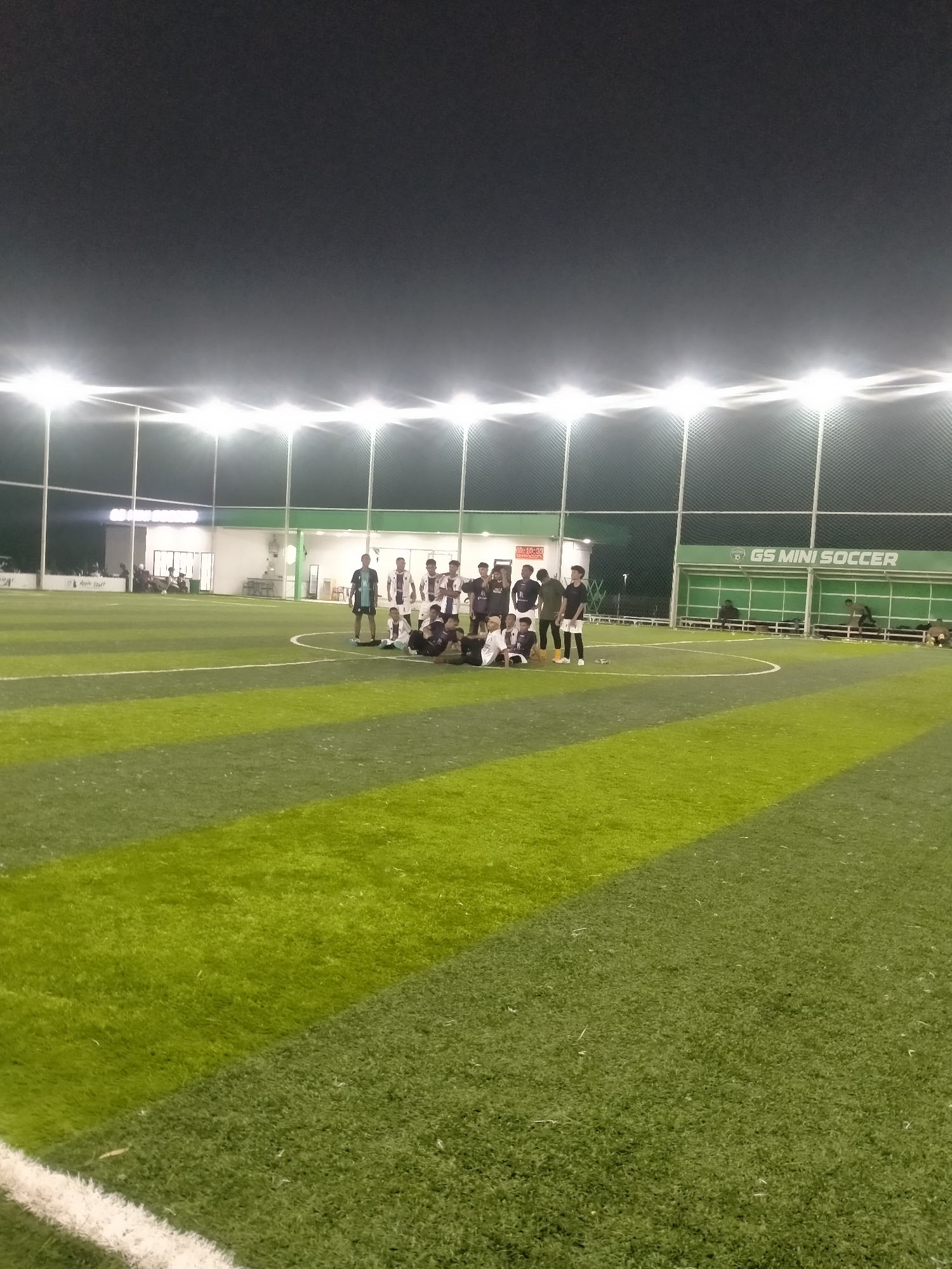 The image captures a group of approximately 15 people, likely a soccer team, gathered at the center of a well-lit, artificial turf soccer field. The field is marked with white lines and varying shades of green turf. Overhead lights are clearly visible, illuminating the field. In the background, there is a building with the signage "GS MINI SOCCER" visible, along with seating areas adjacent to the field. The overall environment indicates an evening soccer match or practice session. The image was taken in Sigli, Indonesia.