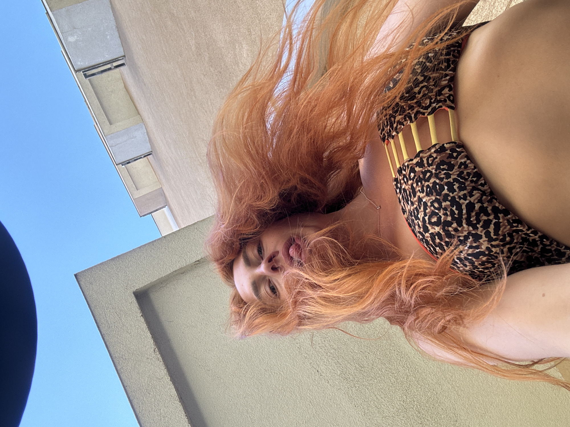 This is a low-angle, close-up, outdoor, self-portrait photograph of a person with long, wavy, red hair. They are wearing a leopard-print bikini with yellow accents. The background consists of a light-colored building and a bright blue sky. The person's face is visible, and they appear to be looking towards the camera with their tongue slightly extended. The lighting suggests it is daytime. The image is oriented sideways.