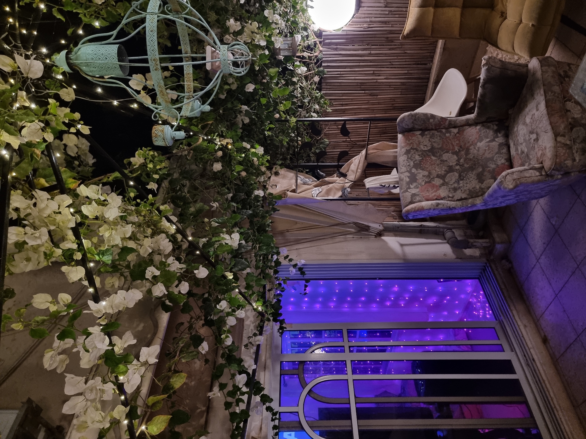 The image shows an interior space with an eclectic mix of furniture and decorations. A vintage armchair with a floral pattern sits beside a modern white plastic chair, with a tan cushion visible in the background. A teal-colored birdcage-style lantern hangs near a wall covered in white flowers and greenery, accented by small string lights. Below, a window with a grid pattern is illuminated by a purple light source, hinting at an outdoor scene. The ceiling is covered in a woven material, possibly bamboo. Given the location of Bat Yam, Israel, the scene likely represents a café, restaurant, or private patio area with a bohemian aesthetic. No people are visible.