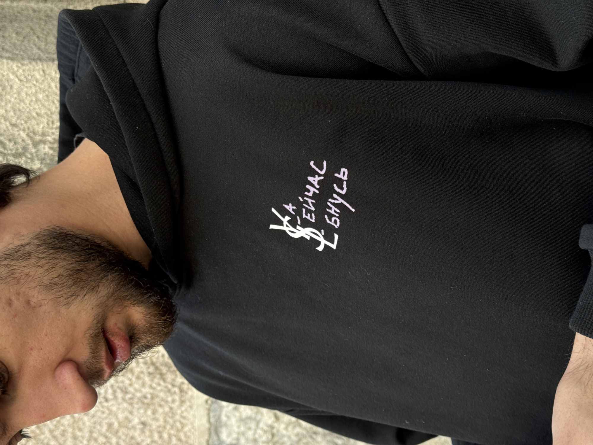 The image displays a close-up, slightly tilted view of a man from his chest upwards, wearing a black hoodie. His face is visible in profile on the left side, showing a portion of his forehead, eye, nose, mouth, and a dark, well-groomed beard. His lips are slightly parted.
The central object is the black hoodie, made of a textured, possibly cotton, fabric. On the left chest area, there's a distinctive design. It features a white, stylized symbol resembling the interlocking "YSL" logo, positioned to the left of two lines of Cyrillic text. The text is written in a light pink or lavender color, in a handwritten style. The upper line reads "ЕЙЧАС" (pronounced "Sey-chas"), which translates to "NOW" in English. The lower line reads "БНУС" (pronounced "Bnuss").
The man is not engaged in any particular activity other than wearing the garment. The background is a blurred, light-colored, textured surface, possibly concrete or pavement, suggesting an outdoor setting during daytime. The location context is Belgrade, Serbia, though no specific visual cues within the image confirm this. The overall impression is casual and focused on the garment's design.