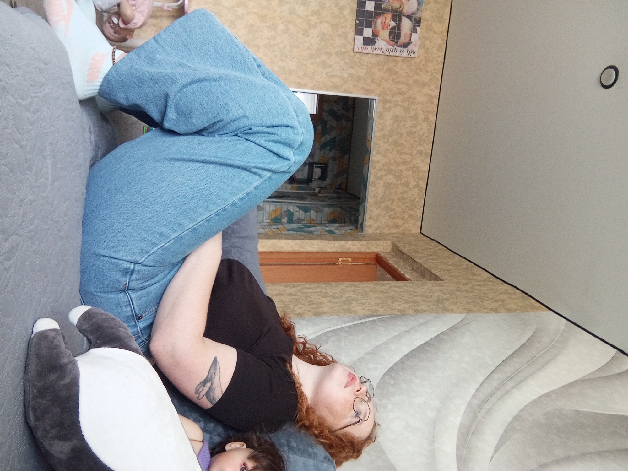 The image depicts a person lying on a grey sofa, wearing a black shirt, blue jeans, and glasses. They have red hair and a tattoo on their arm. A plush toy, white and grey, is near them. A child with purple clothing is partially visible beside the toy. In the background is a wall with patterned wallpaper, a mirror reflecting a tiled room, and a poster. The location is possibly a residence in Yurgamysh, Russia, suggested by the presence of elements associated with a domestic environment.