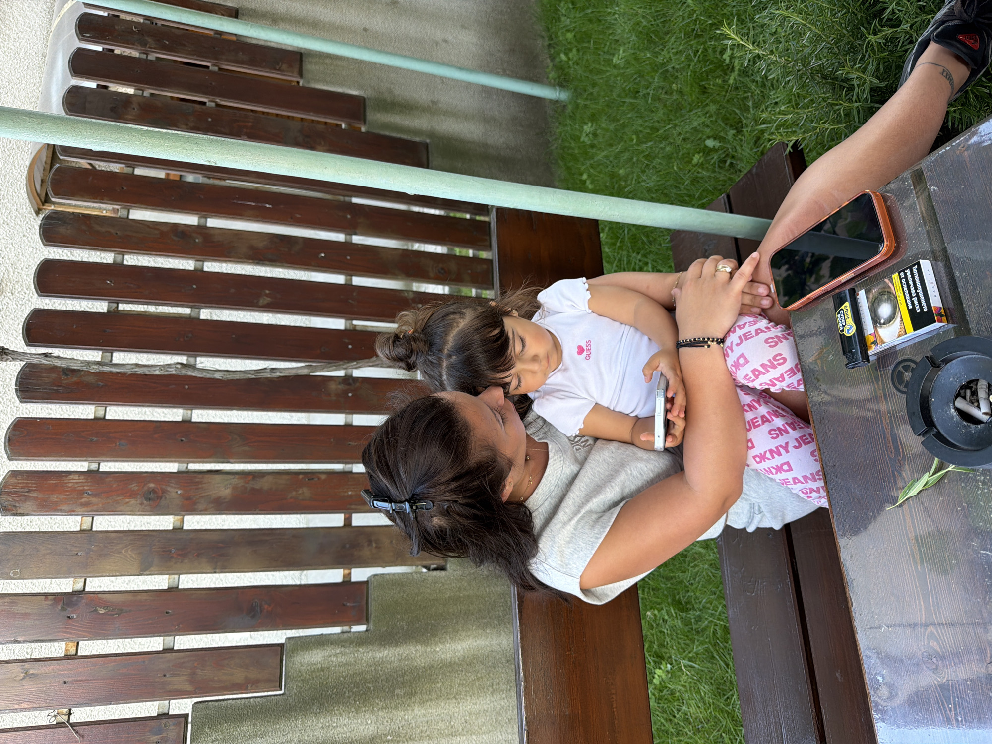 A mother and her young daughter are seated on a wooden bench in a backyard setting. The mother, with dark hair pulled back, is holding the daughter on her lap and looking down at a smartphone. The daughter, wearing a white t-shirt and pink patterned leggings, is also looking at the phone. A table in front of them has a pack of cigarettes and a cell phone on it. The background consists of a wooden fence and lush green grass. The image does not provide any specific visual cues to pinpoint a location in the city of Burgas, Bulgaria.