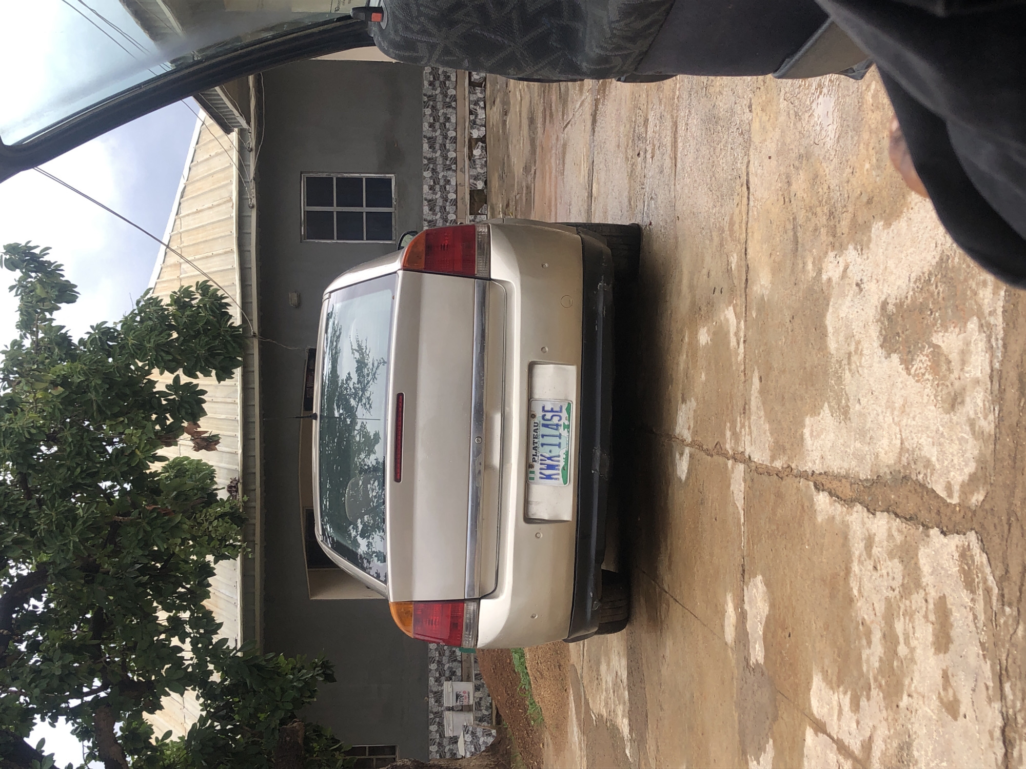 A light-colored sedan is parked in an outdoor concrete area that appears wet, suggesting recent rain. The car, a hatchback, is viewed from the rear, with its license plate reading "KWX-114SE PLATEAU". The background features a building with a metal roof and a large tree with green foliage overhangs the scene. A portion of another vehicle, possibly a van, is visible on the right side of the frame, showing a dark, patterned seat. The ground is a mixture of concrete slabs and dirt, with several puddles of water. The environment suggests a residential or semi-rural setting in New Stadium, Nigeria.