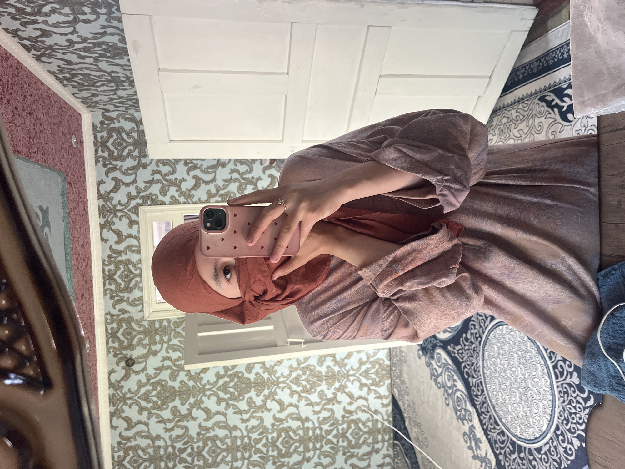 A young person is taking a mirror selfie indoors, possibly in a bedroom. They are wearing a rust-colored hijab and a long, patterned dress in shades of pink and brown. The dress has voluminous sleeves. Their face is partially obscured by the hijab and their hand, which is holding a pink smartphone with heart decorations. They are wearing a ring on their right ring finger. The background shows patterned wallpaper, a white door, and a bed with a decorative rug. The ceiling has an ornate, reflective surface. The environment appears to be a domestic setting in the city of Tashlak, Kyrgyzstan.