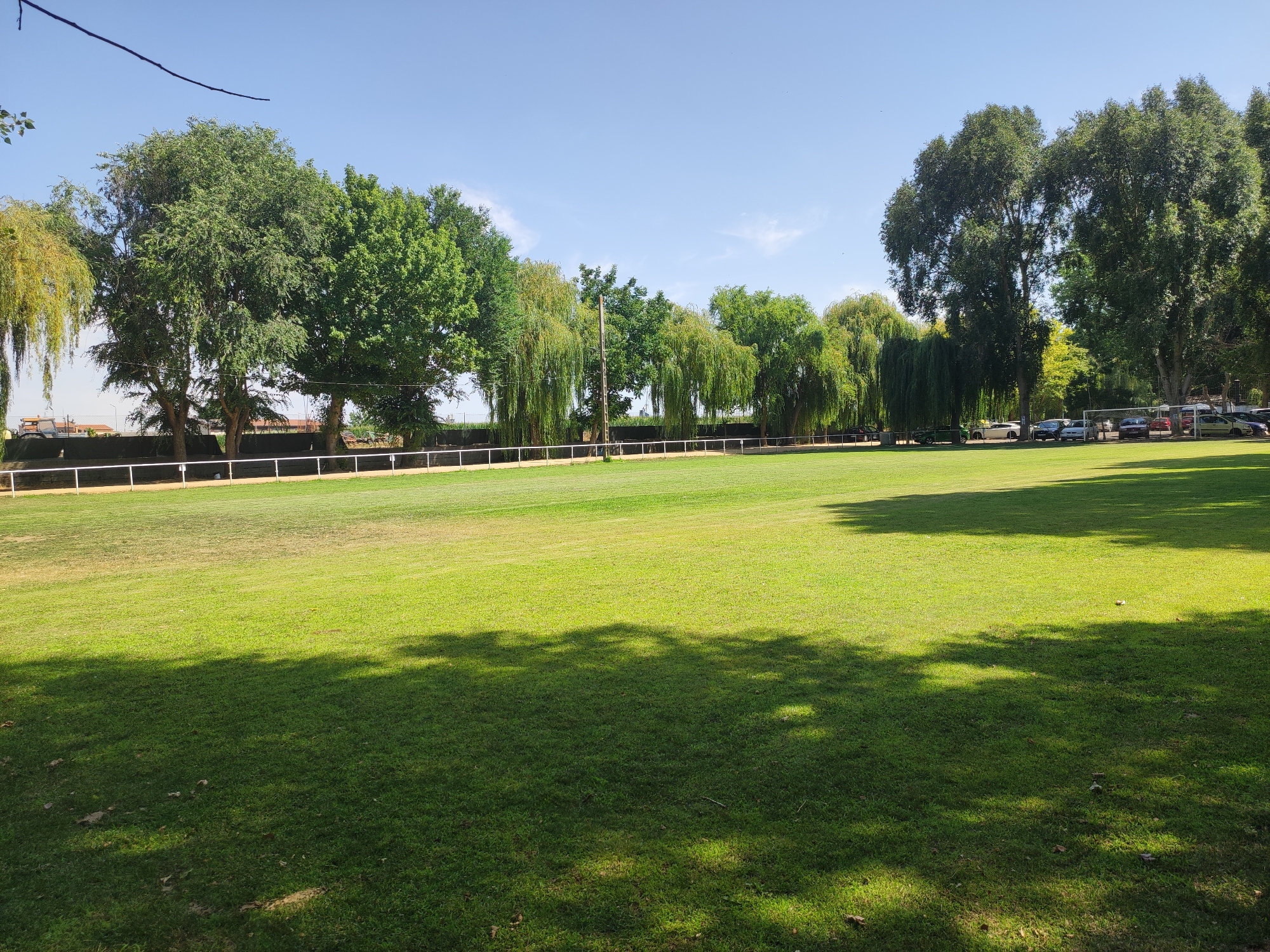 The image shows a grassy field with trees and a clear blue sky. In the background, there is a white fence and several cars parked. The field is well-maintained and appears to be a recreational area, possibly a park or sports field. The sun is shining, casting shadows from the trees onto the grass. The location is identified as Huerta, Spain. The scene is tranquil and bathed in natural light.