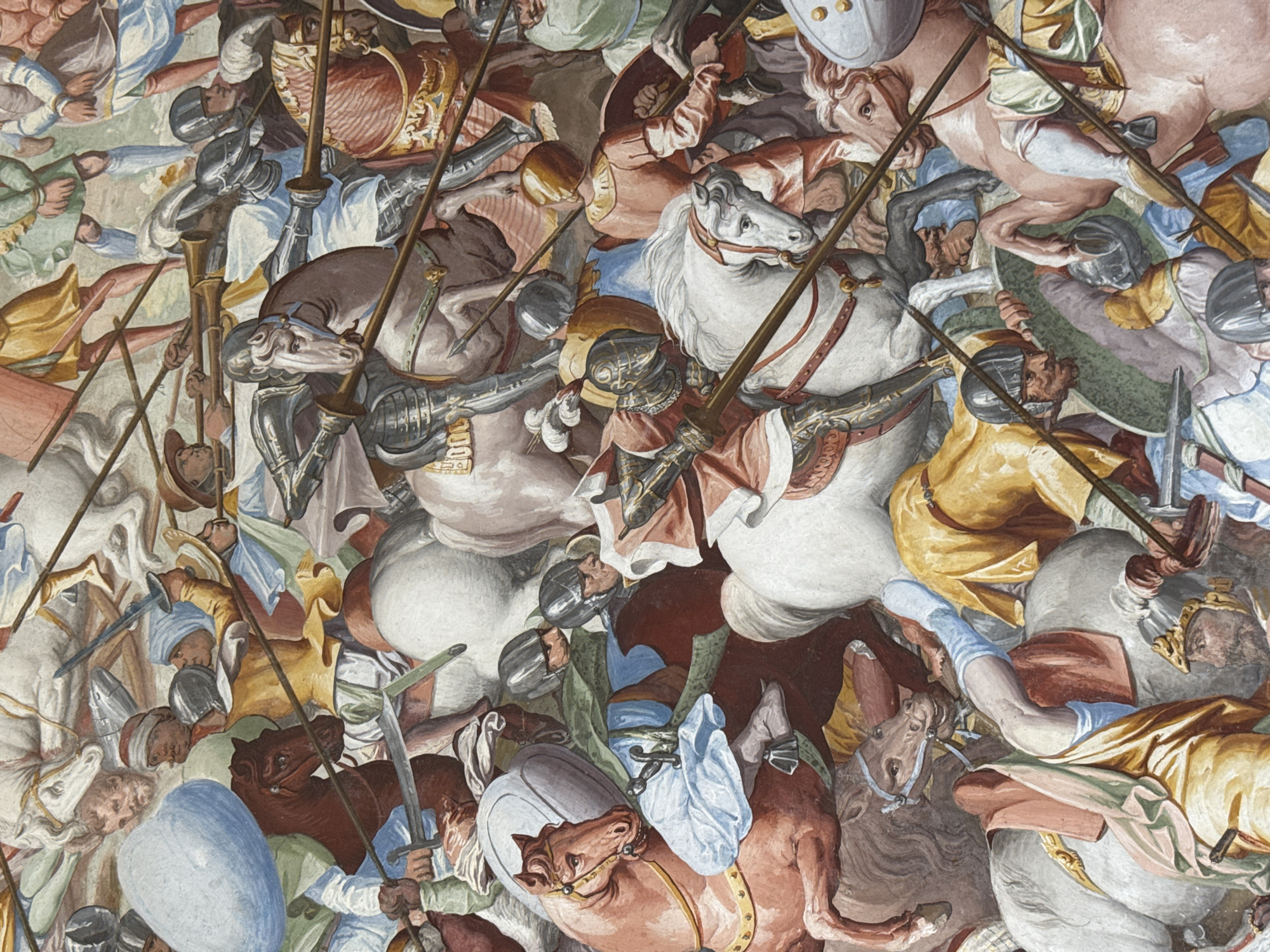 This is a fresco painting depicting a chaotic battle scene with many figures on horseback. The subjects are soldiers in historical armor and clothing, wielding spears, swords, and shields. Several horses are prominently featured, some white, others brown and grey. The composition is dense, with figures overlapping and creating a sense of intense action. The color palette includes muted tones of browns, greys, whites, and reds, with accents of blue and yellow.
The event captured is a fierce cavalry engagement. Soldiers are shown lunging with their weapons, and the horses appear to be in full gallop, adding to the dynamism of the scene. The expressions on the faces of the soldiers are difficult to discern due to the scale and style of the painting, but the overall impression is one of intense conflict and struggle.
Notable details include the ornate armor worn by some of the riders and the various styles of helmets and shields. The fresco appears to be a ceiling or wall painting, given its perspective and the way the figures are arranged. There are no visible cues for time of day or weather. The setting is an interior space, likely a grand hall or chapel, suggested by the fresco style and historical context of San Lorenzo De El Escorial, Spain. No visible text is present in the image.