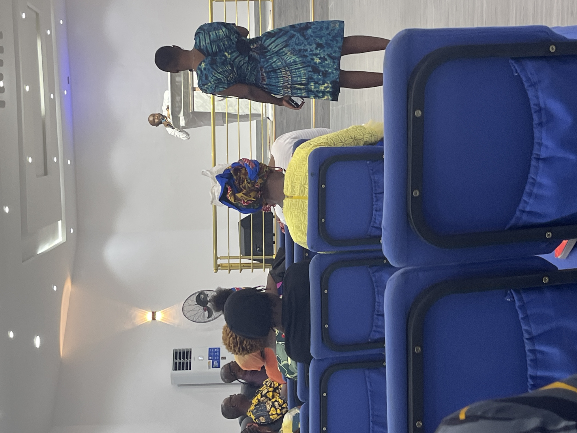 The image shows the interior of a building in Ado, Nigeria, likely a church or gathering place, populated with individuals seated in blue chairs. A woman in a patterned blue dress stands in the foreground, holding a phone. Several rows of people are visible. A man in a white shirt is visible on a balcony speaking into a microphone. An air conditioner is mounted on the wall along with an electric fan. The walls are white.