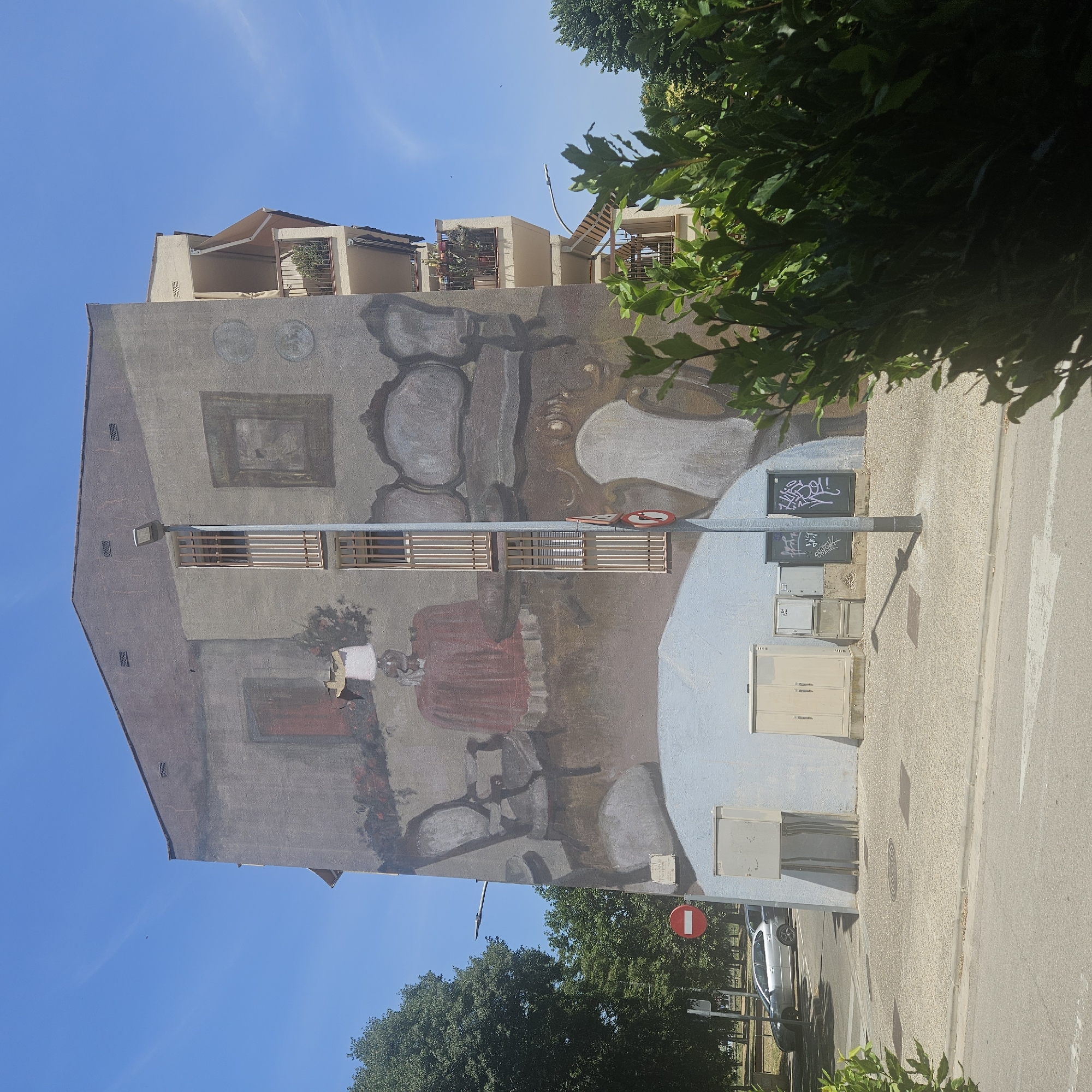 The image depicts a mural painted on the side of a residential building located in the city of Girona, Spain. The mural is an optical illusion, creating the appearance of an interior scene of a room. Furniture, such as chairs and a table with a red tablecloth, are depicted. A lamp sits on the table, and framed artwork hangs on the wall. Two balconies with plants are visible on the right side of the building. The lower part of the building has several utility boxes and electrical conduits. In front of the building, there is a sidewalk, a street, and a parked silver car. Trees and greenery surround the building and the street. The sky is clear and blue. A red and white circular sign is visible on a pole.