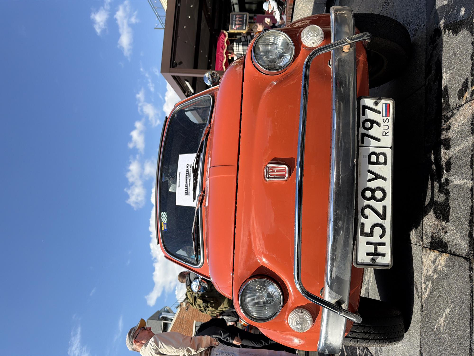 A low-angle, front-oriented outdoor shot depicts an orange vintage car bearing a Russian license plate with the registration number "H528YB 797 RUS." The car features round headlights, chrome bumpers, and a rectangular emblem on the front. A white paper is affixed to the windshield with text in Russian. The car is parked on a paved surface of square stone tiles. In the background, blurred figures of people can be seen. The sky is visible with scattered white clouds. The scene is located in Moscow, Russia.