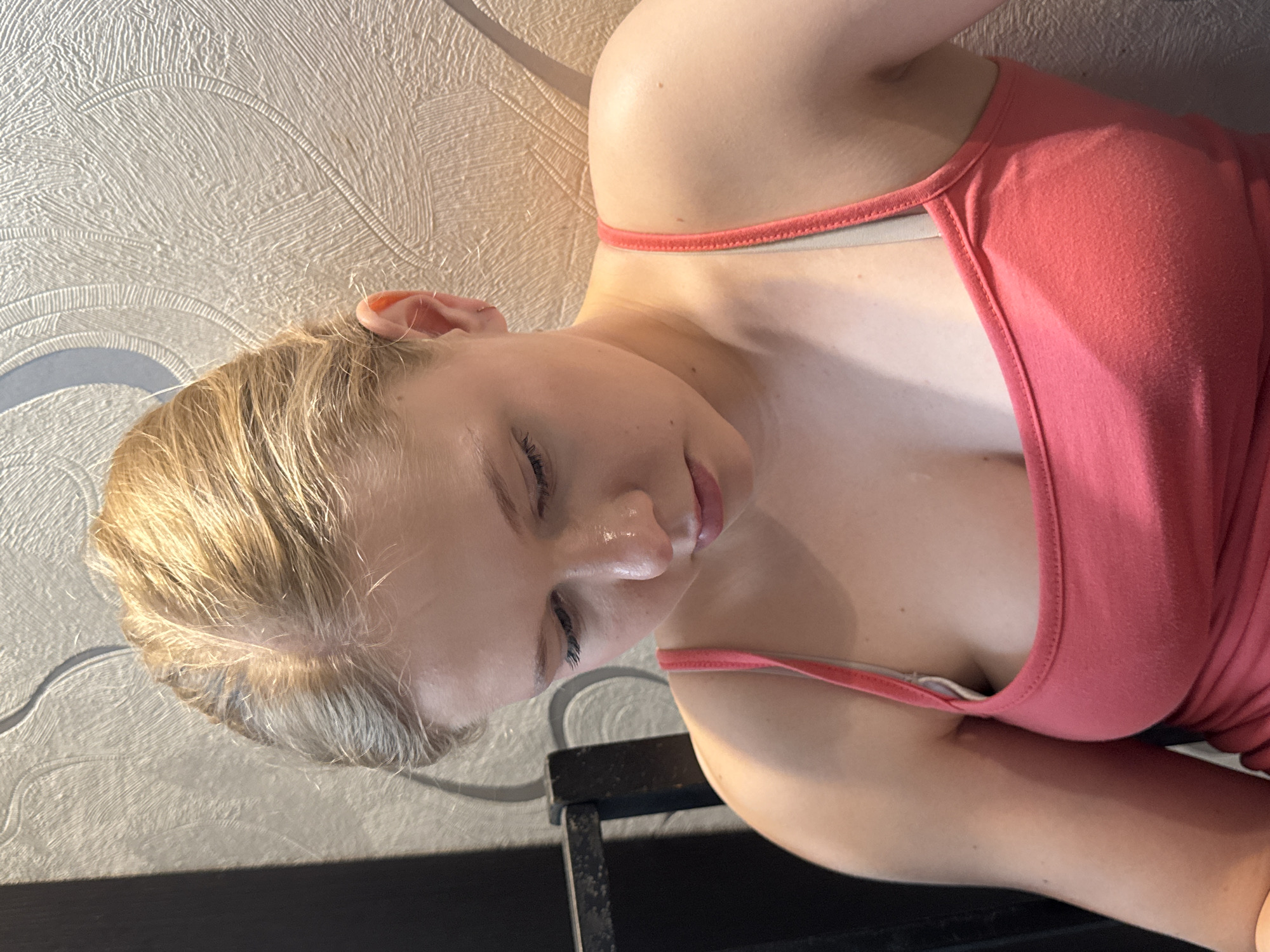 The image depicts a young, fair-skinned female with blonde hair pulled back, gazing downwards and slightly to her left. Her expression appears pensive or focused. Prominent highlights on her forehead and nose suggest direct lighting. She is wearing a coral-pink sleeveless tank top with thin straps; a light gray bra strap is visible beneath the left tank top strap. A small, thin, silver-colored hoop earring is visible in her right ear. The background consists of a light-colored wall with textured, patterned wallpaper featuring an abstract, curvilinear design. To the far left, a dark, vertical object, likely part of a chair or piece of furniture, is visible, casting a distinct shadow. The overall scene is illuminated, with a strong light source appearing to originate from the upper right, creating defined highlights and shadows. This scene is located in the city of Perm, Russia.
