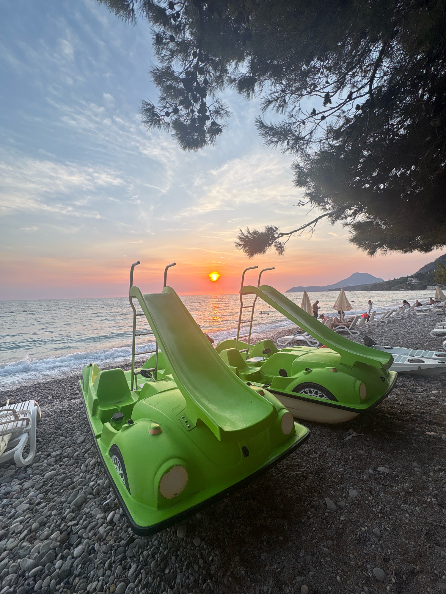 The image depicts a serene beach scene at sunset in Bar, Montenegro. Two bright green pedal boats, each equipped with a slide, are parked on a pebble beach in the foreground. One boat is closer to the viewer, angled towards the right, while the second boat is positioned behind and to the right of the first. The sea stretches out to the horizon, with gentle waves lapping the shore. The sky is a canvas of warm colors, transitioning from soft pinks and oranges near the setting sun to cooler blues overhead, with scattered clouds.
In the background, the shoreline is dotted with beach chairs and umbrellas, suggesting a public beach area. Several people are visible, some lounging on chairs and others walking along the beach. Mountains form a distant backdrop, silhouetted against the colorful sky. The scene is framed by the dark branches of a pine tree with cones, casting a partial shadow over the top of the image. The overall atmosphere is tranquil, capturing the end of a day at the beach.