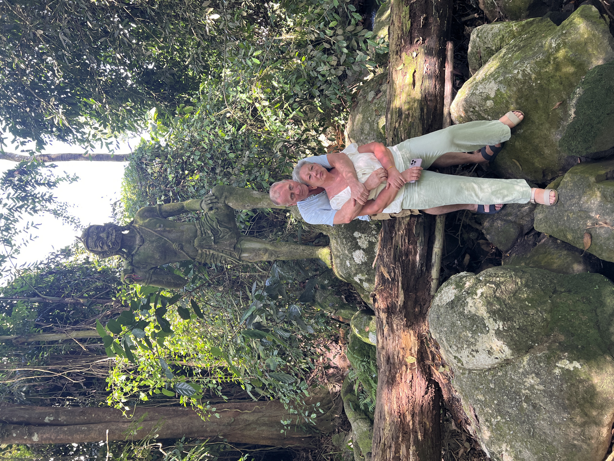 An outdoor, medium shot captures an elderly Caucasian couple posing in front of a moss-covered statue in a forest environment, possibly in Ban Bang Khao, Thailand. The man, wearing a light blue short-sleeved button-down shirt and tan shorts, stands behind the woman, embracing her from behind. He has his arms wrapped around her waist and his head leaning slightly on her shoulder. The woman, with gray hair and wearing a white sleeveless blouse and light green wide-leg pants, faces the camera with her hands clasped in front of her. She is positioned in front of the man and slightly to the right, leaning her head towards him. She is holding a mobile phone in her left hand. Both are barefoot. The statue, depicting a hunched figure with a stylized face and wearing a loincloth, is positioned behind the couple and to the left, and appears to be made of stone, covered in moss. The surroundings are lush and green with dense foliage, large trees, moss-covered rocks, and a large fallen tree trunk in the foreground. The lighting suggests it is daytime, with sunlight filtering through the leaves, creating dappled shadows. The overall scene evokes a sense of nature and ancient structures.