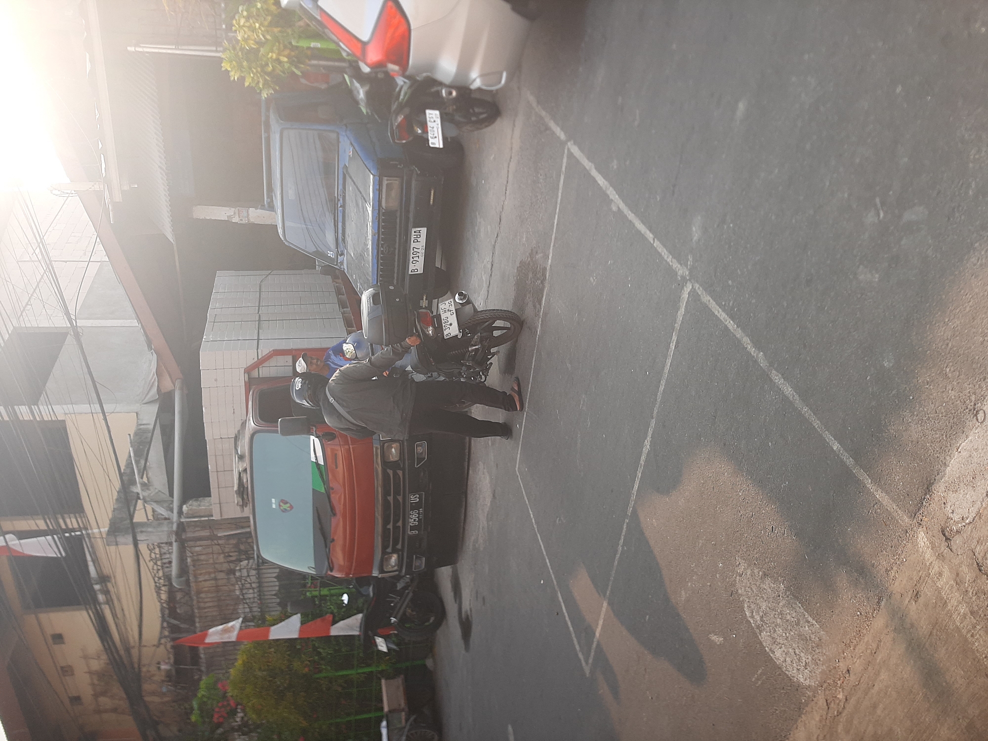 The image depicts an outdoor scene in Central Jakarta, Indonesia, likely a street or a parking area, during daylight hours with bright sunlight creating lens flare. In the foreground, a person is standing next to a motorcycle. The person is wearing a dark jacket and pants, and a helmet. The motorcycle is dark-colored with a top case. Behind them, a blue SUV is parked. To the left of the SUV, a red truck is also parked, loaded with white rectangular objects, possibly bricks or building materials. Another person is visible inside the truck, wearing a white cap. Several other motorcycles are parked in the vicinity. The ground is paved asphalt with white lines marking parking spaces. The background shows buildings and some greenery.