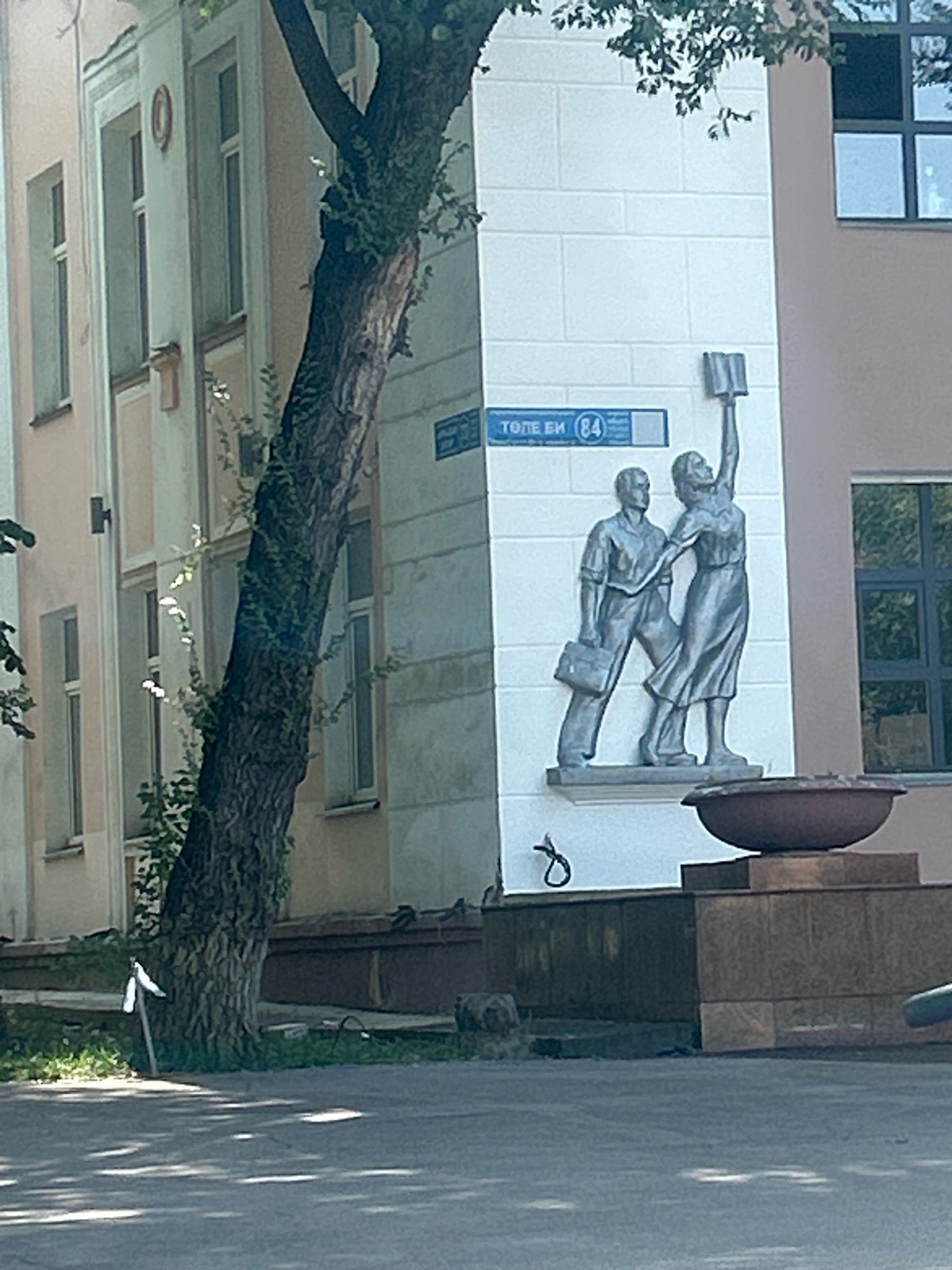 The image shows a building facade in Almaty, Kazakhstan, with a relief sculpture of two figures. The sculpture depicts a man in a suit holding a briefcase and a woman reaching up to hold a book. A street sign above the sculpture reads "ТОЛЕ БИ (84)", indicating Tole Bi Street and house number 84. A large tree with dark bark and green leaves partially obscures the building. The building is light-colored with multiple windows. In the foreground, there is a paved surface, likely a sidewalk or road, with shadows cast from the tree. A large, dark-colored, bowl-shaped object sits on a raised platform to the right of the sculpture. The overall scene appears to be an outdoor urban environment on a sunny day.