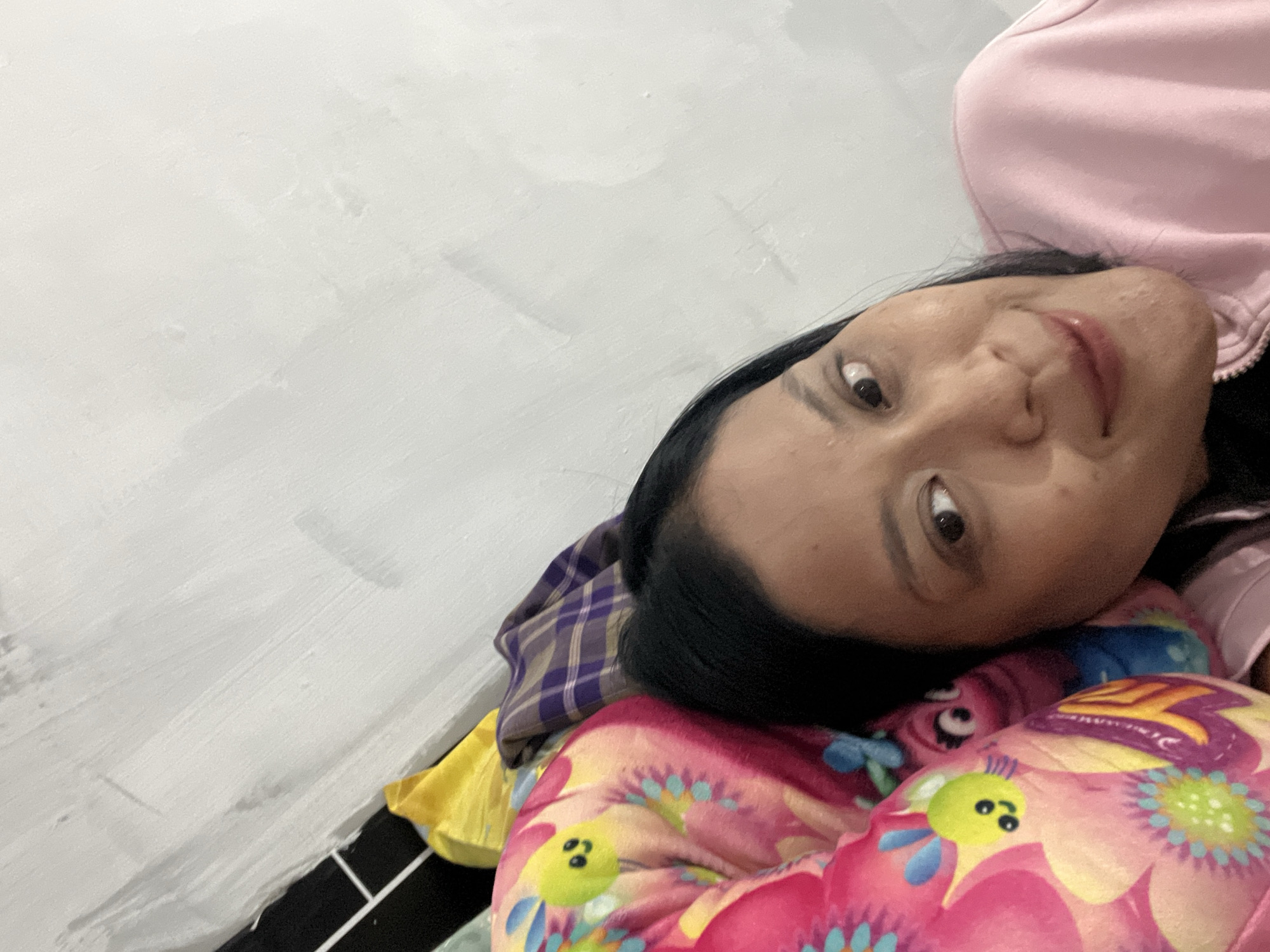 A woman is lying down next to a pillow with a cartoon character on it. She is wearing a pink jacket. The background is a wall with white paint.
