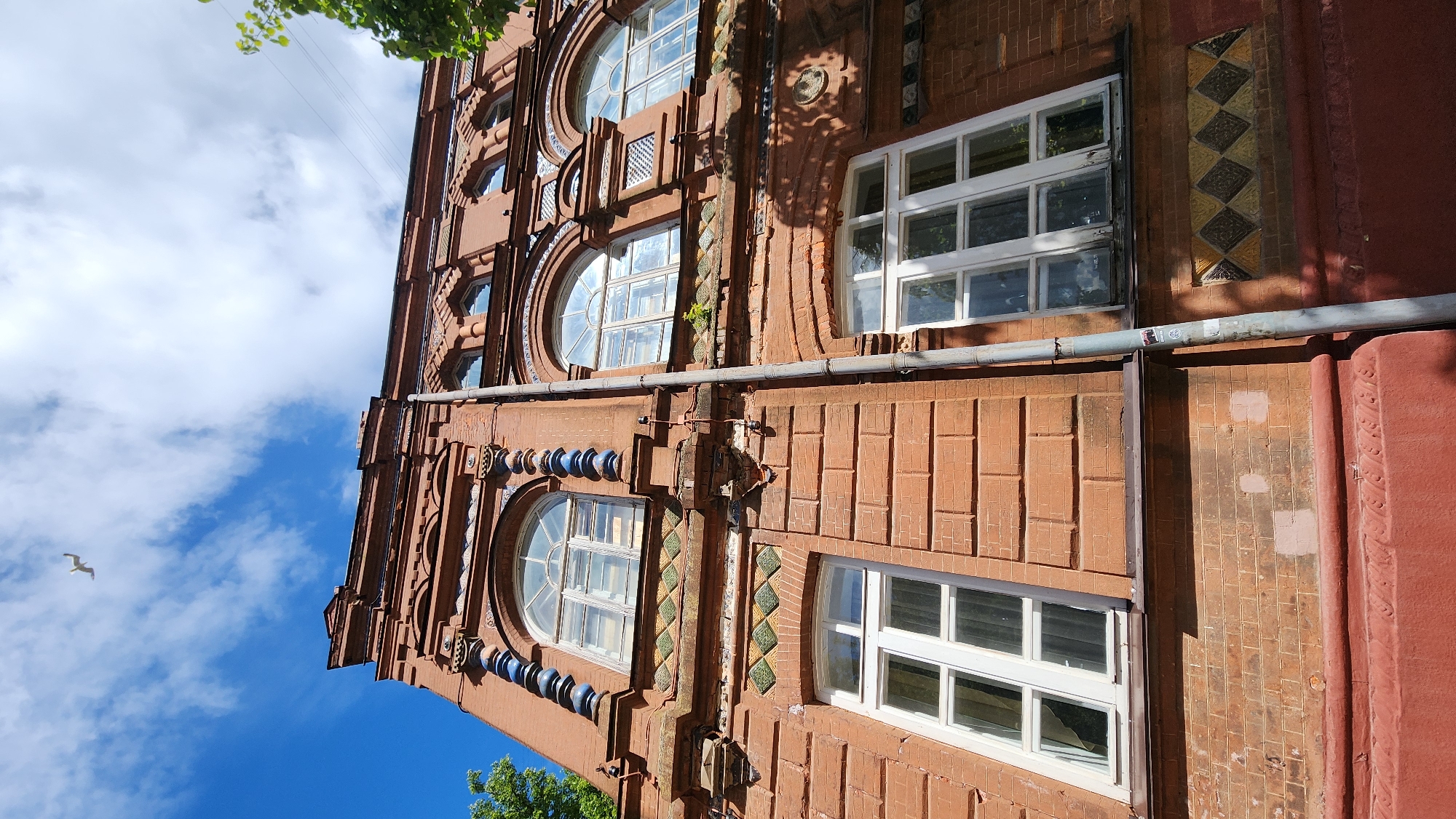 A low-angle, eye-level shot depicts a brick building with a gray metal pipe running horizontally across the façade. The building's architecture features circular windows and rectangular windows with white frames. Decorative tiles with diamond patterns are visible between the windows. The building is painted in shades of red and brown. The sky is blue with scattered white clouds, and a single seagull is flying in the upper left corner of the frame. The precise location in the city of Unknown, Unknown is not clear from the provided image.