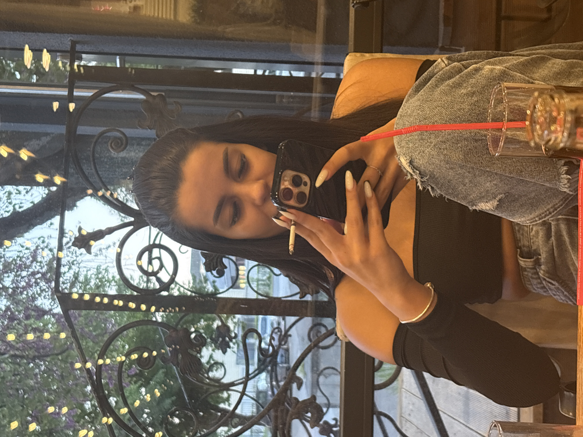 A young woman sits at a table in Plovdiv, Bulgaria. She wears a black top and denim jeans, and has long dark hair. She is holding a cell phone to her face while also holding a lit cigarette. A glass with a red straw sits on her lap. Outside the window is a wrought-iron fence with lights. Trees and buildings are visible in the background.