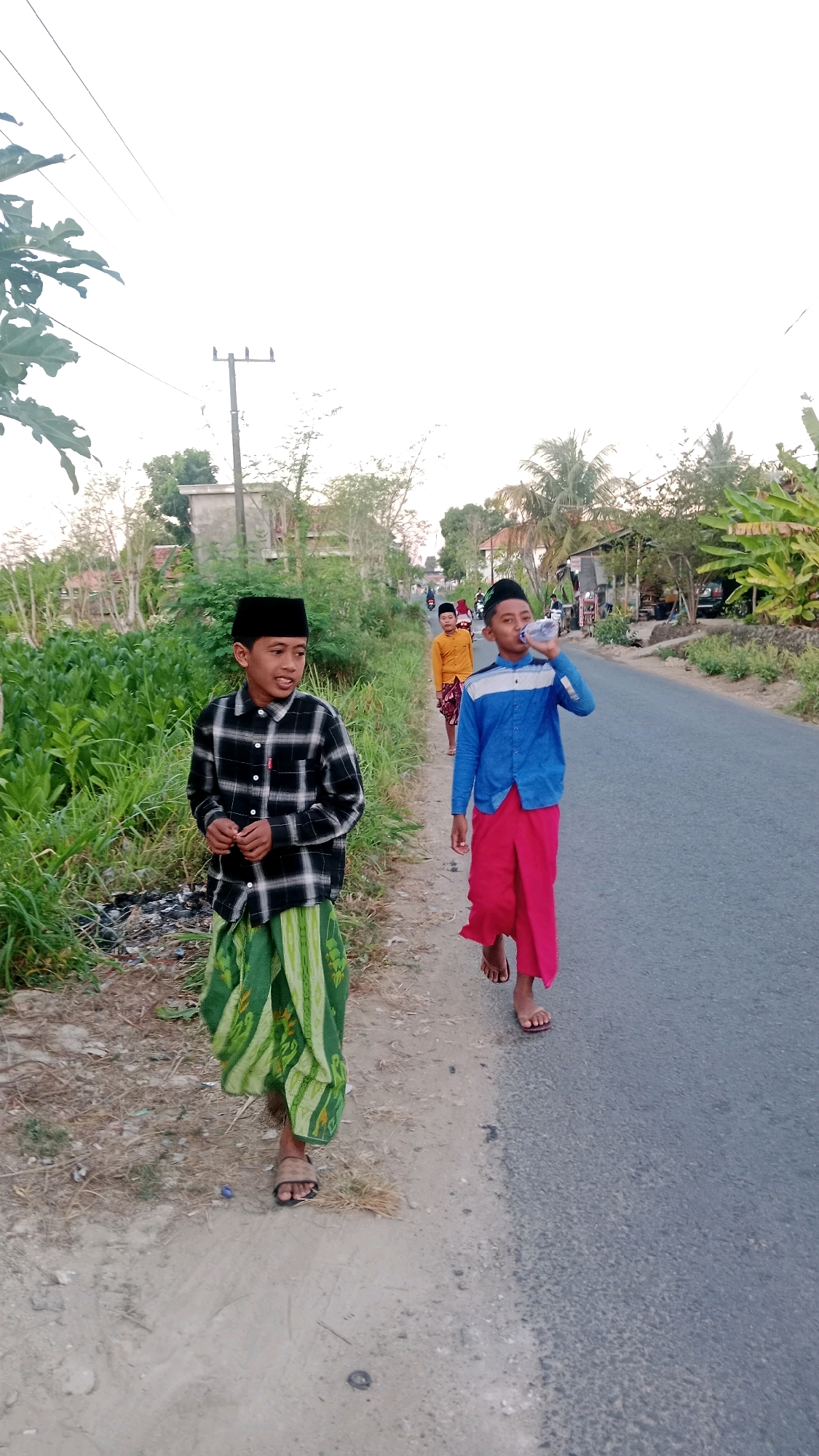 A medium shot captures four young males walking along a rural road in Dempo Barat, Indonesia. The perspective is from eye level, slightly angled downwards towards the road. The primary subjects are two boys in the foreground. The boy on the left, facing slightly towards the camera, wears a black and white checkered long-sleeved shirt, a black skullcap, and a green sarong with a patterned design. He is holding something small in his right hand. To his right and slightly behind, another boy is walking on the road, facing away from the camera and drinking from a clear plastic bottle. He is wearing a blue long-sleeved shirt with white and blue stripes on the collar and cuffs, a black skullcap, and bright pink pants. Further down the road, two more boys are visible in the background, one wearing a yellow shirt and a patterned sarong, and the other partially obscured. The road is paved and asphalted, with a dirt and gravel shoulder to the left, lined with lush green vegetation. In the distance, scattered houses and trees suggest a rural or semi-urban setting. The sky is overcast, with a pale, hazy light. The overall scene depicts a casual outdoor stroll, likely during daylight hours, with the boys dressed in traditional Indonesian attire.
