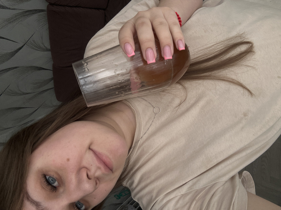 A female subject, primarily seen from the chest up, is depicted in an indoor setting within the city of Chaikovsky, Russia. She has light skin, blue eyes, and light brown hair partially framing her face. Her lips are slightly parted in a faint smile, and a small mole is visible on her right cheek. She is wearing a plain, light beige short-sleeved t-shirt with several faint, dark smudge marks on the front. A thin, silver-colored necklace with a small circular pendant rests on her neck. Her left hand, adorned with fingernails featuring a French manicure with bright pink tips, holds a clear, stemless glass containing a brownish, amber-colored liquid. The liquid level suggests partial consumption, and some residue is present on the inner surface of the glass. A red beaded bracelet is visible on her left wrist. The background features a dark gray wall covered with wallpaper exhibiting a subtle pattern of light gray, abstract, swirling lines. A portion of dark brown or burgundy upholstered furniture, possibly a sofa or armchair, is visible to the right of the subject. The lighting is diffused, casting soft shadows.