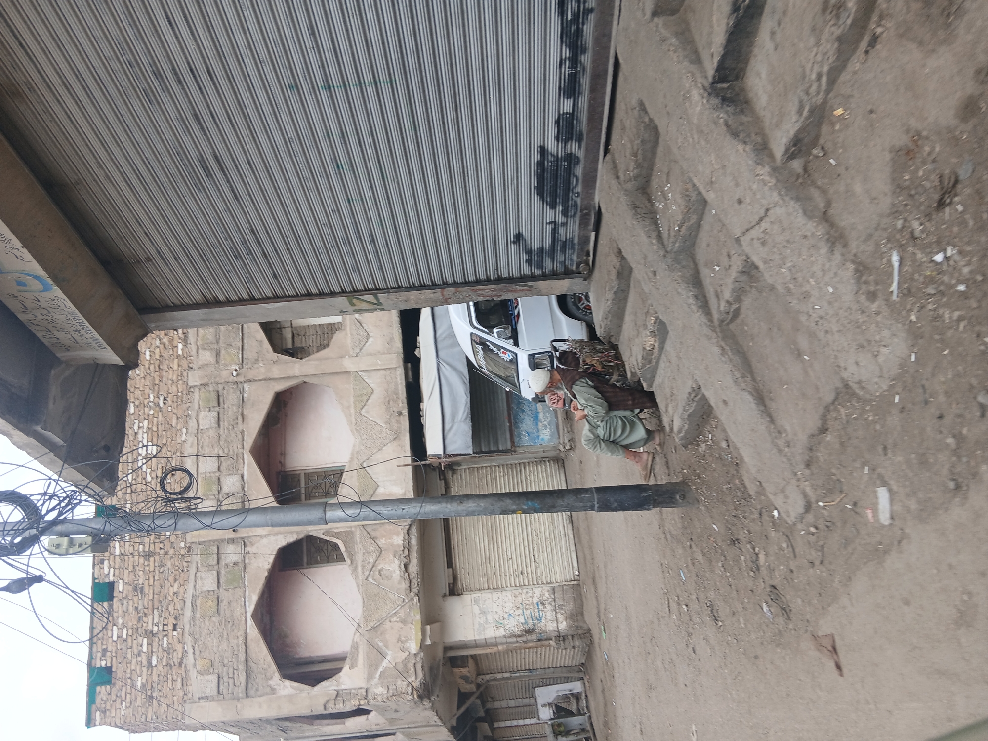 A low-angle, medium-shot photograph depicts a street scene in Quetta City Tehsil, Pakistan. An elderly man, dressed in traditional attire, is squatting on the sidewalk with a book in his hands. A white vehicle is partially visible behind him. Buildings line the street, with one building featuring unique, honeycomb-shaped window insets. Overhead, a network of electrical wires crisscrosses the frame. The sidewalk and street are dirt. A closed metal shutter suggests a business is closed. The scene is in daylight.