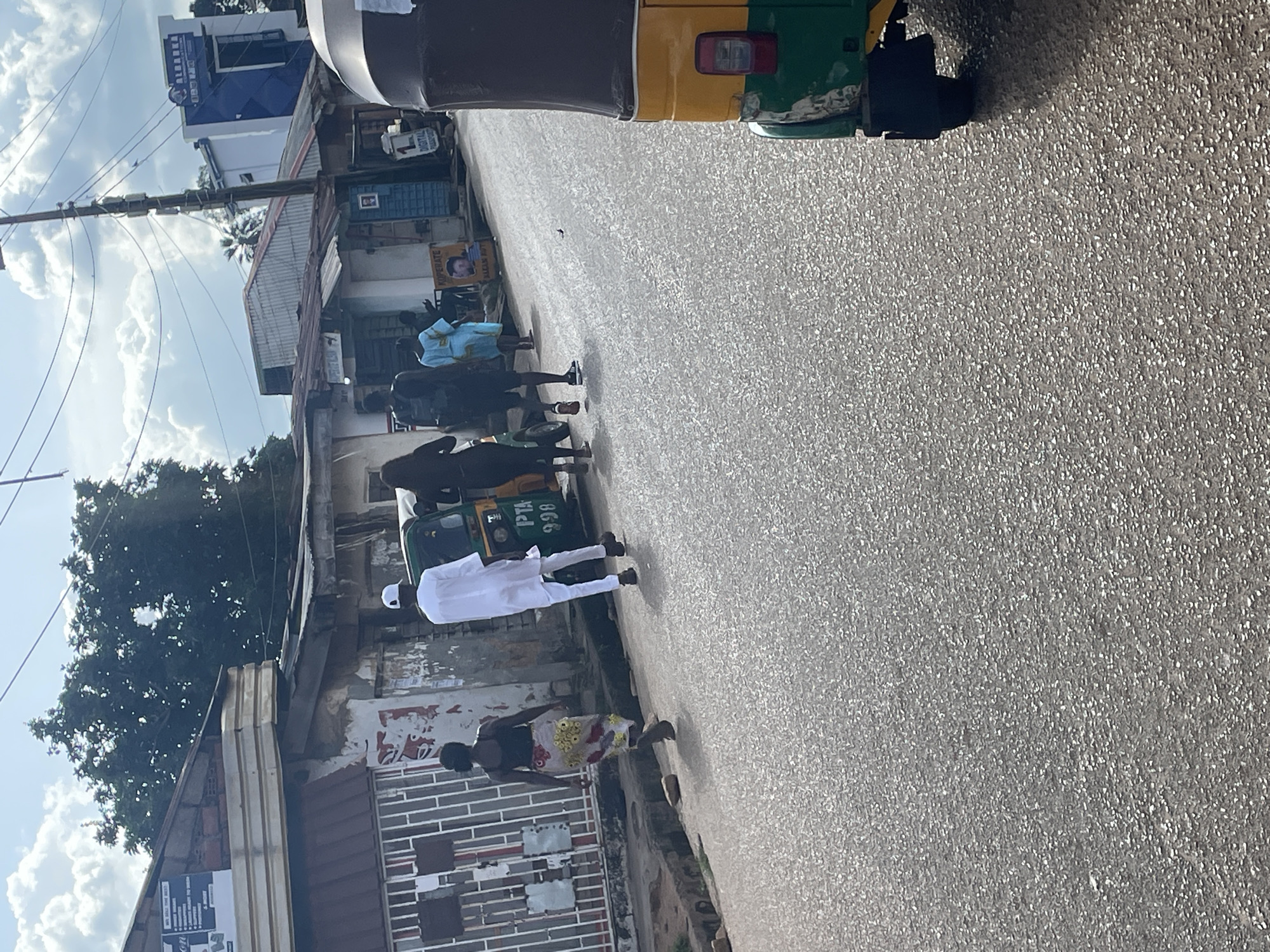 The image depicts a street scene in Jos, Nigeria, under a cloudy sky. In the foreground, the right side of a green and yellow auto-rickshaw is visible. The street itself is paved with asphalt, showing texture from the aggregate. On the left side of the street, a woman in a colorful patterned skirt and black top walks away from the camera. Behind her, a man in white traditional attire and a white cap walks towards the right. Further down the street, two other individuals are walking away from the camera, one in dark clothing and the other in a light blue garment. Several buildings line the street, with some showing signs of age and weathering, including corrugated iron roofs and crumbling plaster. One building has a white fence in front of it. Signs are visible on some of the buildings, including one with text that appears to be in English. Electrical poles and wires are present overhead, indicating infrastructure. The overall impression is a bustling, everyday street in an urban environment in Nigeria.