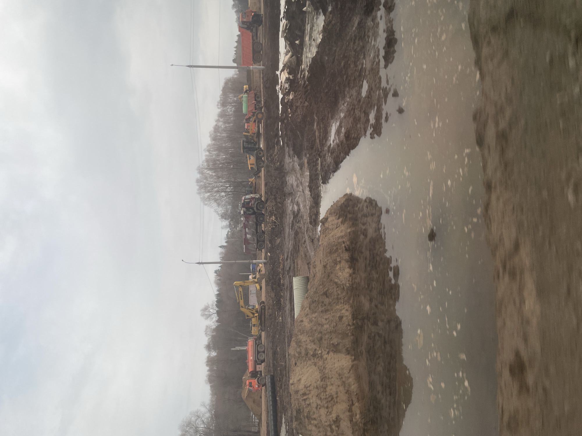 The image depicts a construction site in what appears to be Kabanikha, Russia. There is a large amount of standing water and mud. Heavy equipment, including excavators, dump trucks, and tractors, are visible in the background. Trees with bare branches are scattered throughout the scene. Power lines and a light pole are visible above. The sky is overcast and gray. There are no people visible in the image.