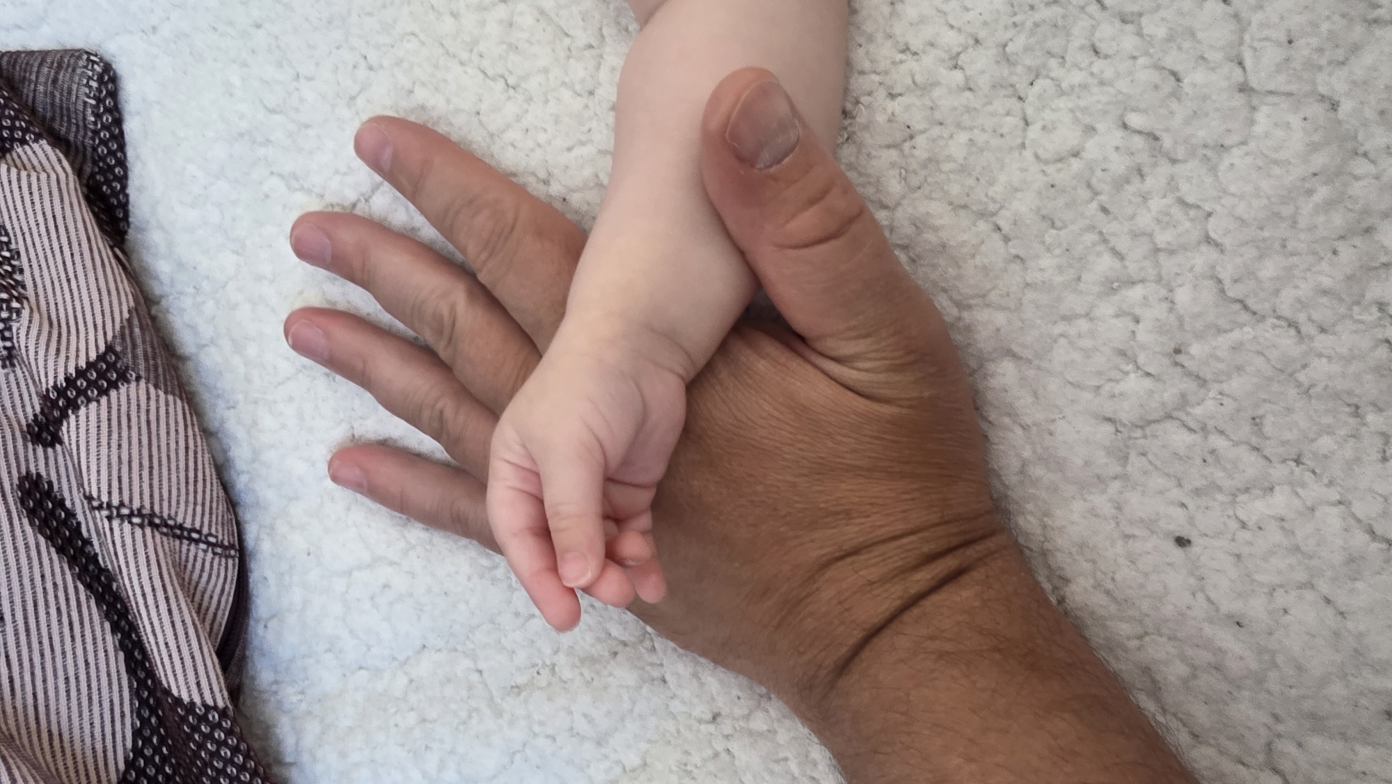 A close-up, overhead shot features an adult's hand holding a baby's hand against a white, textured surface. A patterned cloth lies to the left of the hand. The adult's hand is darker in complexion than the baby's. No location is discernible.