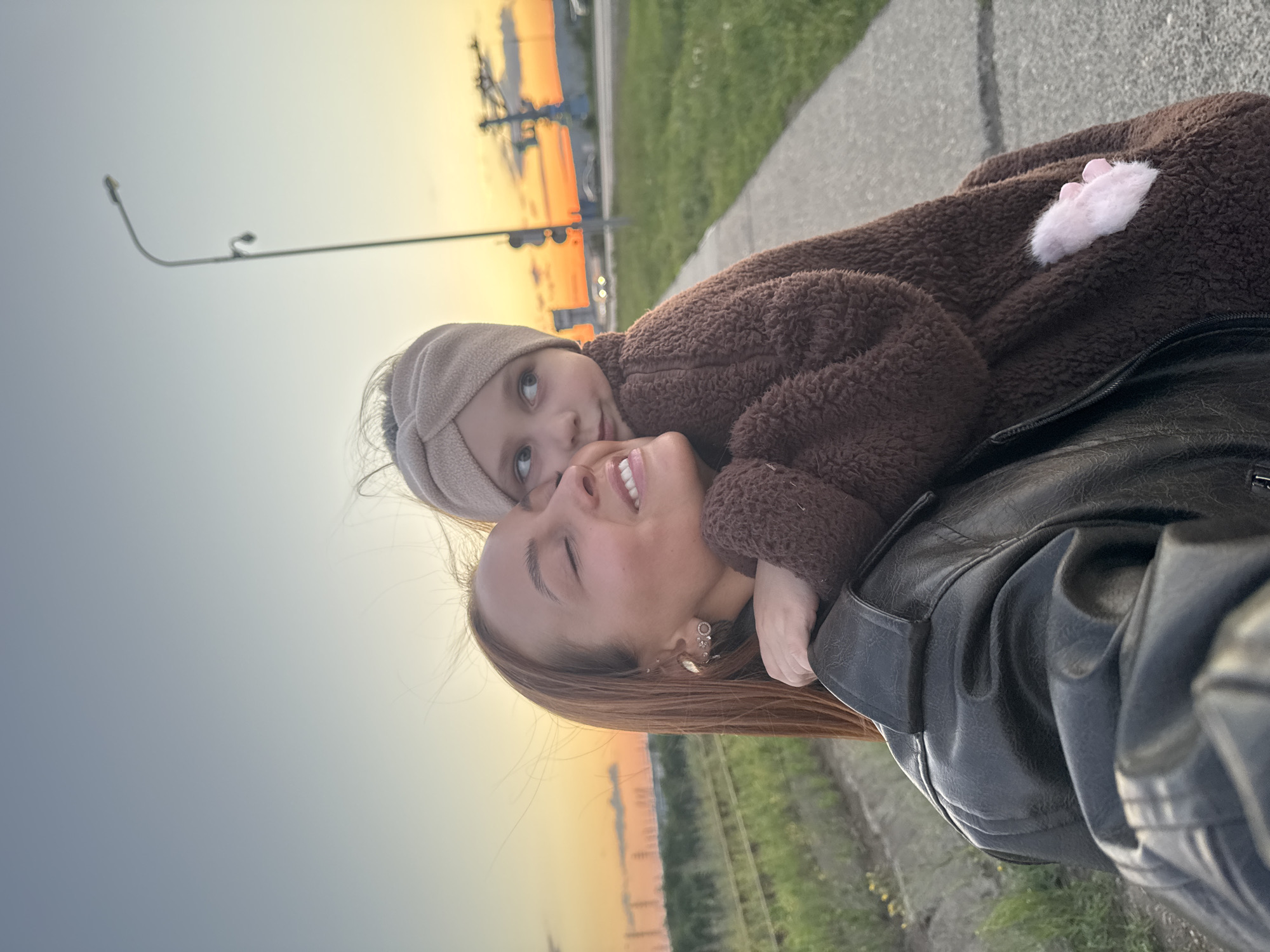 A medium shot shows a woman and a young girl embracing in an outdoor setting during sunset. The woman, positioned in the foreground and slightly to the left, is looking upwards with her eyes closed and a smile on her face. She has long, auburn hair and is wearing a black leather jacket. The girl is behind her, hugging her and looking to the right, with her head resting on the woman's shoulder. The girl is wearing a brown, fuzzy jacket with a beige headband.
The background features a paved pathway, a grassy area, and a glimpse of railroad tracks on the left. In the distance, a vibrant sunset paints the sky with orange and yellow hues, silhouetting industrial structures and a tall lamppost. The scene appears to be located on the outskirts of Chaikovsky, Russia, near the Kama River. The overall impression is one of warmth and familial affection amidst a developing industrial landscape.