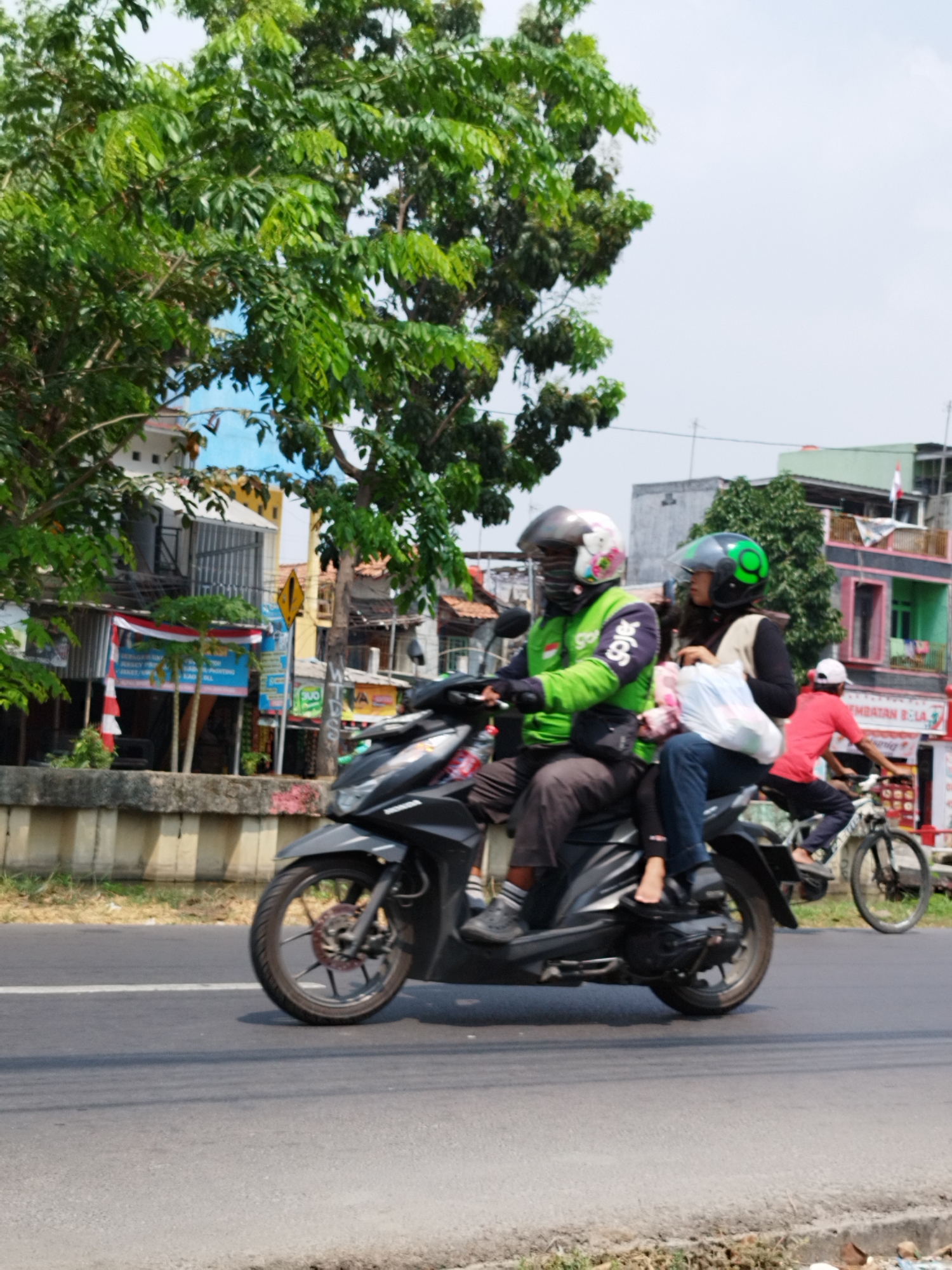 In Bekasi, Indonesia, a man in a green jacket and helmet rides a black motorcycle, with a woman behind him wearing a black jacket and a green helmet. She holds a white plastic bag. A person in a red shirt and white cap rides a bicycle in the background. The scene depicts a sunny day with a cloudy sky, lush green trees, and buildings lining the street. The road appears to be paved, with a concrete barrier along the side. The motorcycle has its headlights on, and the wheels are in motion, suggesting the vehicle is moving. The overall atmosphere is that of a typical street scene in a bustling Indonesian city.