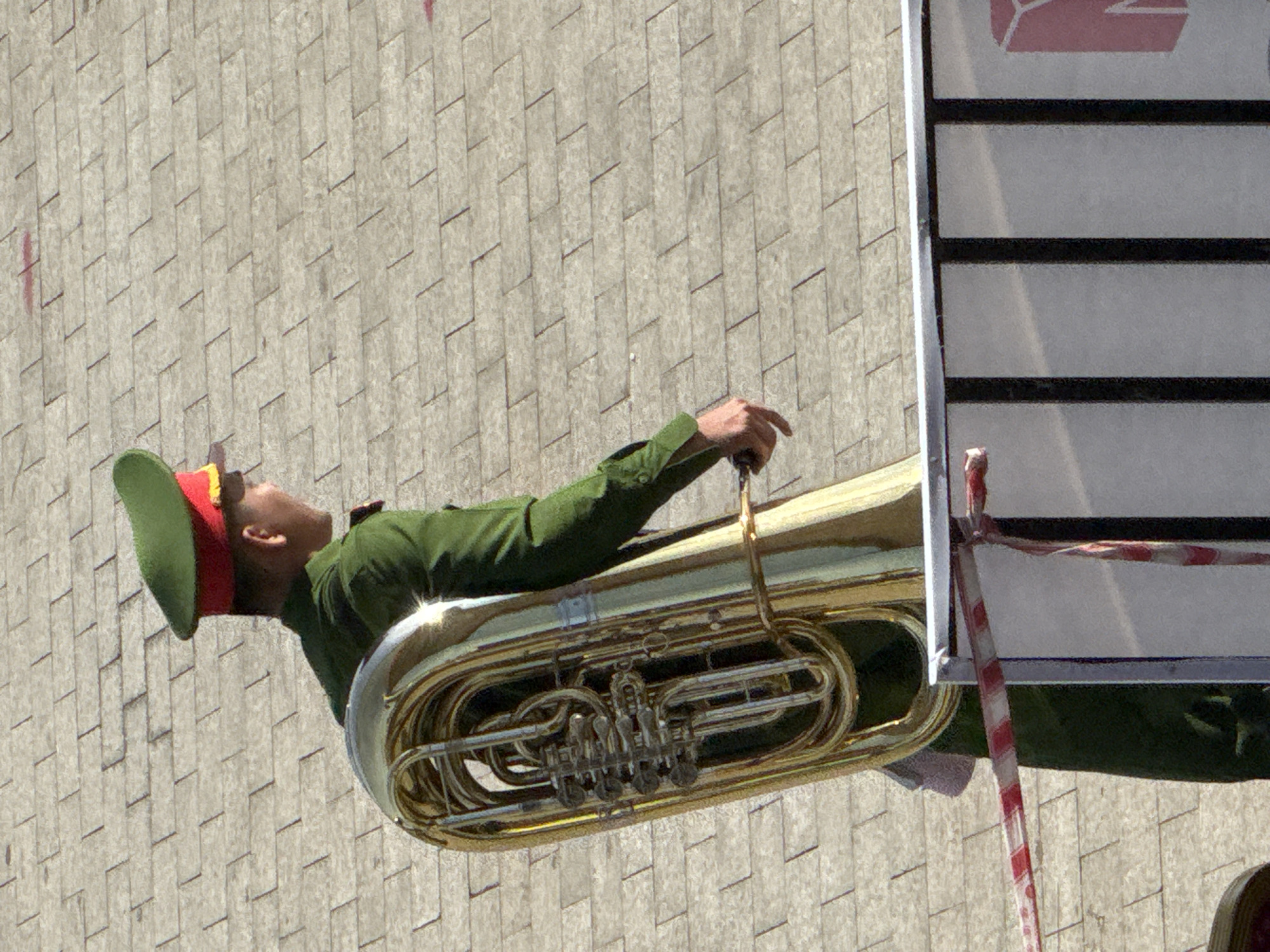 An individual in a green uniform with a red-trimmed hat carries a large brass tuba. The person is walking on a paved surface composed of rectangular grey stones arranged in a brick pattern. A red and white striped tape barrier runs horizontally across the lower portion of the image, in front of a black railing. A portion of a white structure with black vertical bars and a pink graphic element is visible behind the tape. The scene appears to be outdoors, possibly in a public space in Da Lat, Vietnam, given the architectural style and the subject's attire.